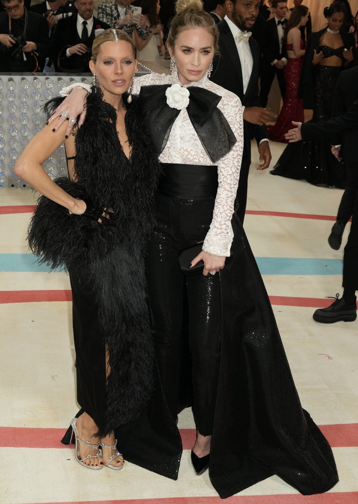 Emily Blunt and Sienna Miller make a striking pair at the 2023 Met Gala, with Blunt in a bold black-and-white ensemble featuring a lace blouse and oversized bow, while Miller stuns in a feathered black gown with silver embellished sandals