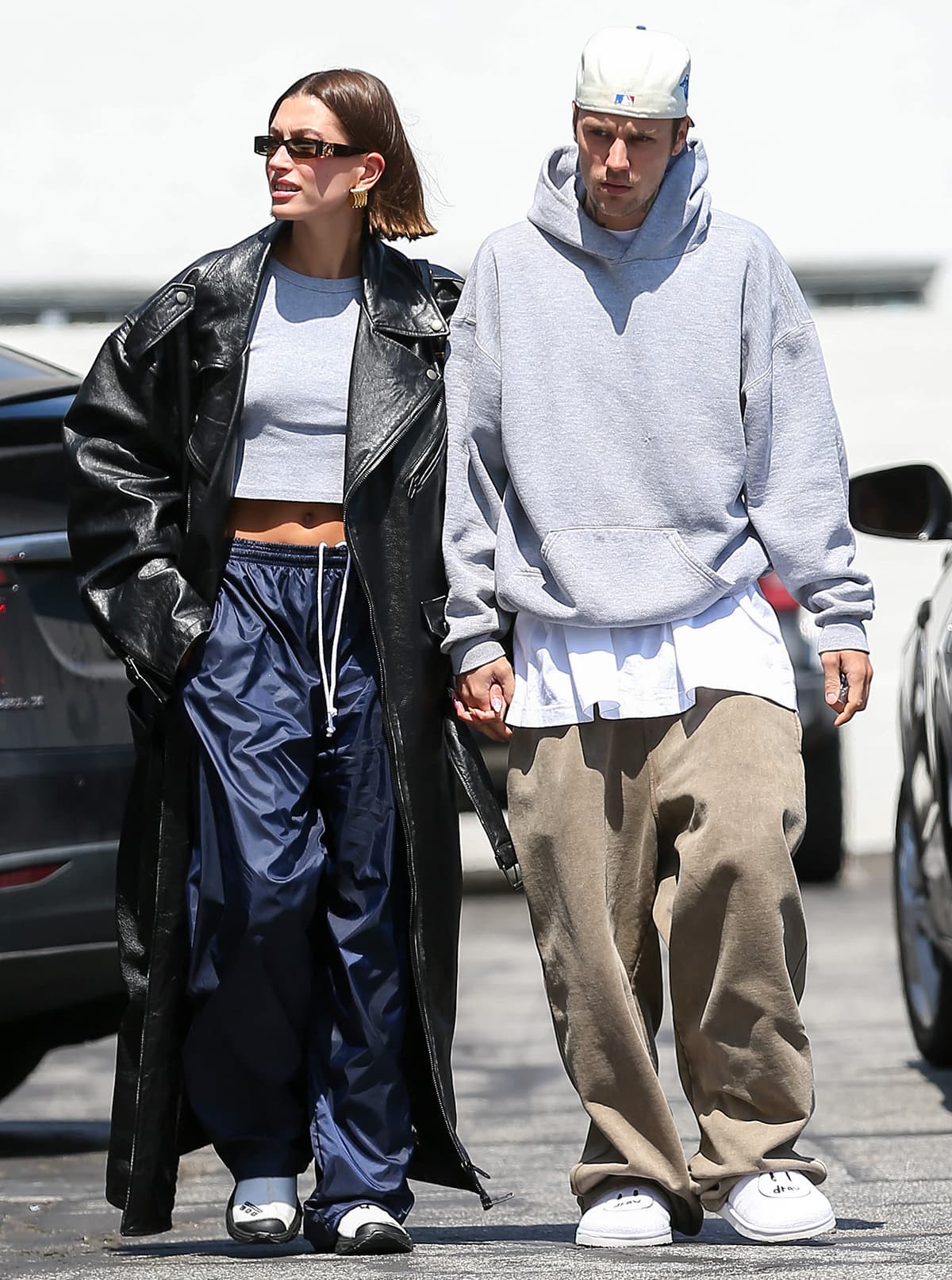 Hailey Bieber flashes her abs in a cropped gray tee, navy parachute pants, and a Balenciaga leather trench, finishing the look with Sandy Liang x Salomon RX Moc 3.0 sneakers, while Justin Bieber keeps it casual in House of Drew slides and slouchy taupe pants