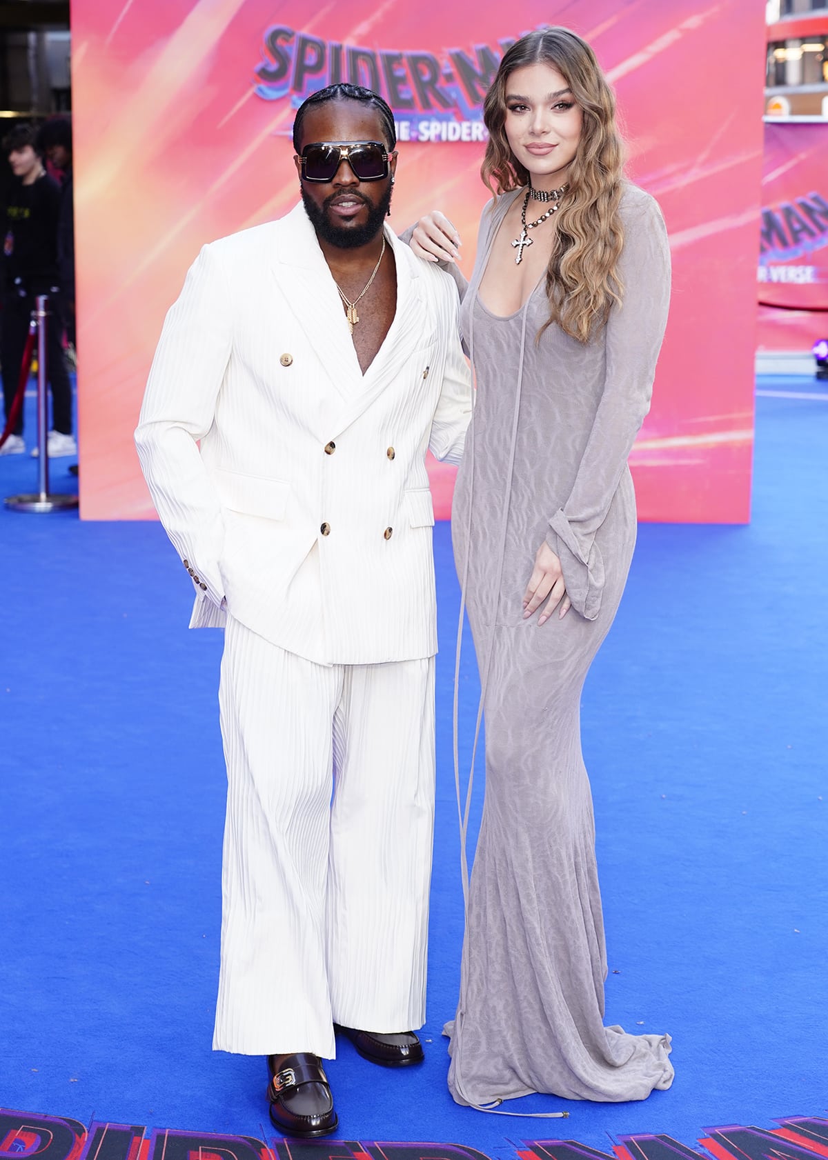 Hailee Steinfeld exudes gothic elegance in a curve-hugging gray Blumarine gown, towering over co-star Shameik Moore, who looks effortlessly dapper in a white ribbed suit with layered gold necklaces and sleek black loafers