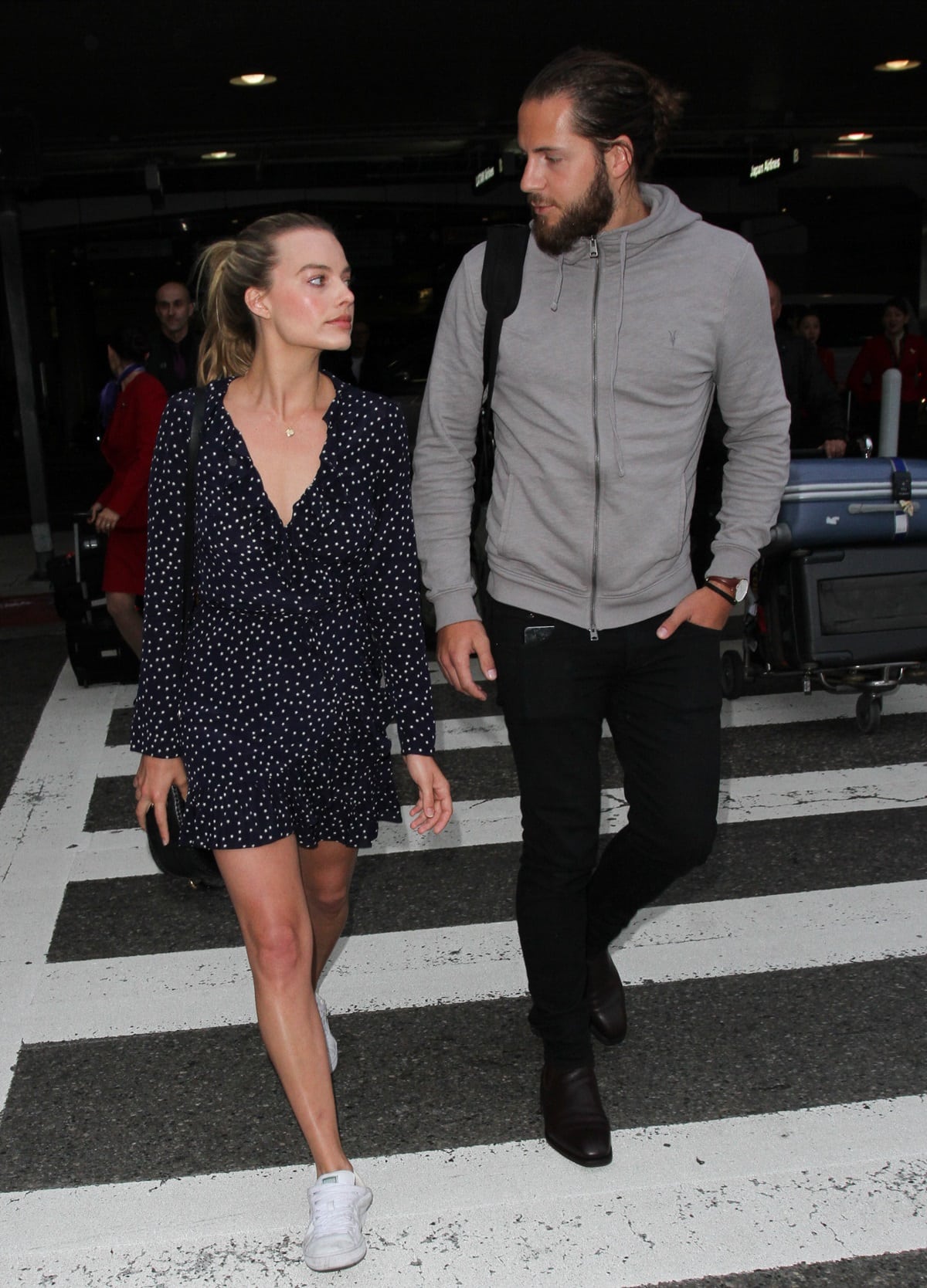 Tom Ackerley, standing much taller, looks down at Margot Robbie as they stroll together, with her in a playful polka dot dress and sneakers, showcasing their height difference