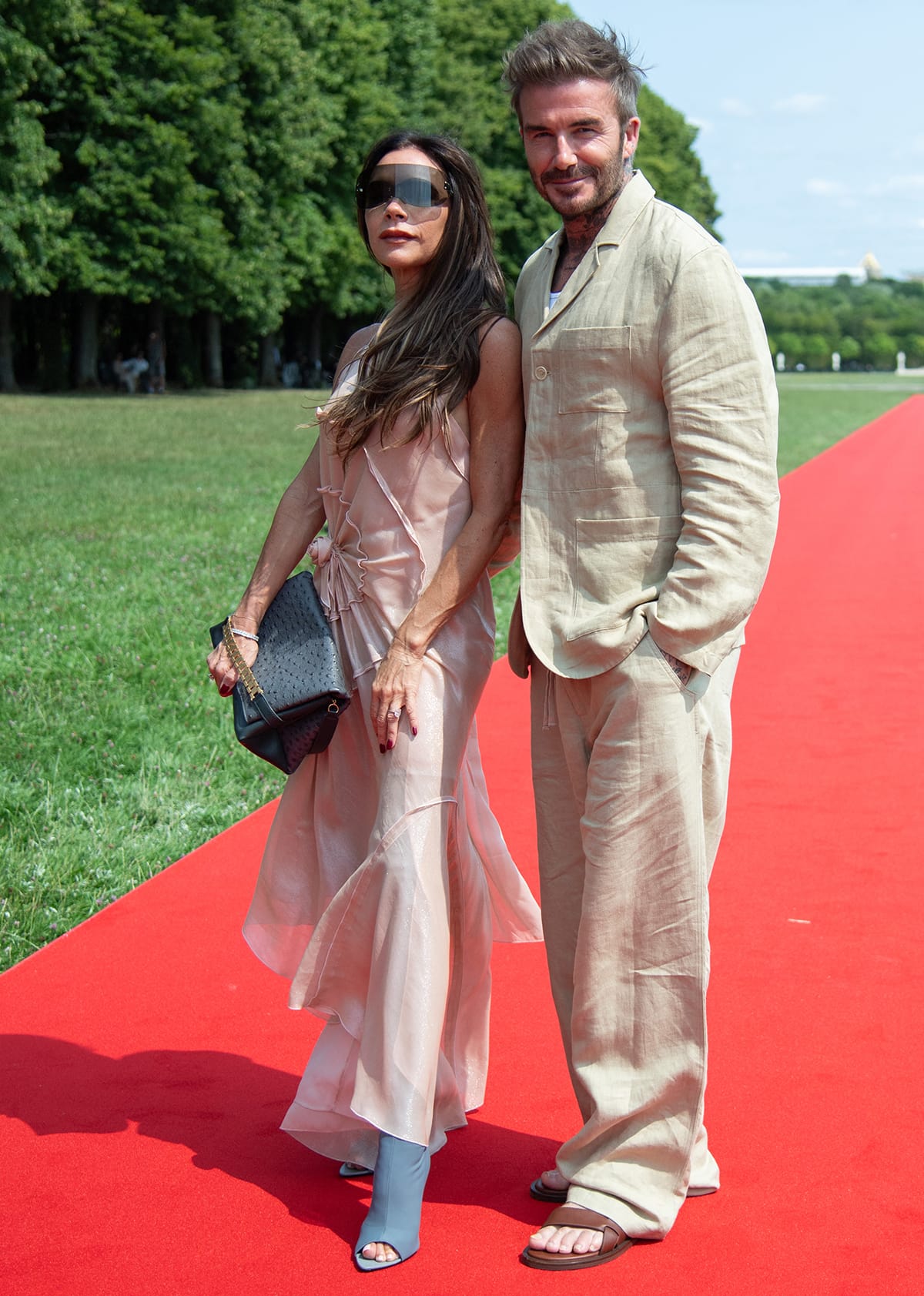 Victoria Beckham and David Beckham pose in coordinating neutral outfits, with Victoria wearing a blush slip dress and thigh-high peep-toe boots