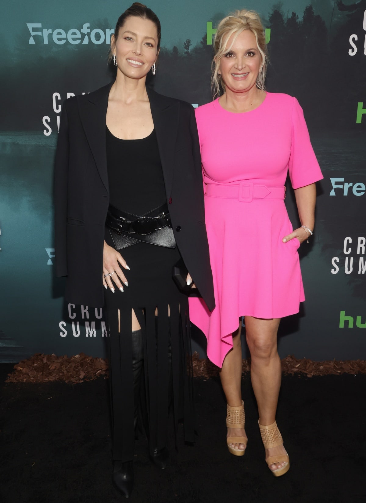 Michelle Purple radiates feminine confidence in a bright pink belted mini dress with asymmetric hemline, paired with woven nude platform sandals that added height and a touch of summery softness next to Jessica Biel’s sharp over-the-knee boots and structured tailoring