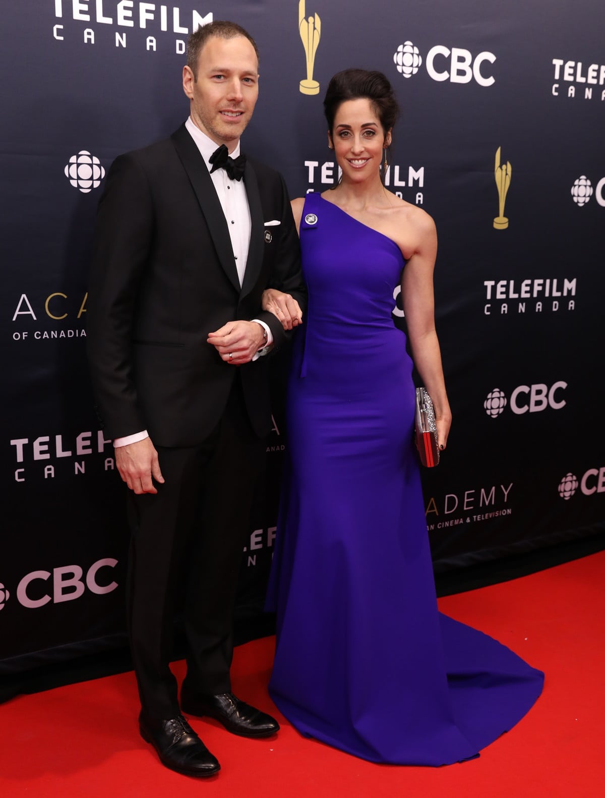 Catherine Reitman dazzles in a one-shoulder royal blue gown with a metallic clutch, while her shoes remain hidden beneath the floor-length hem; husband Philip Sternberg complements her look in a classic black tuxedo at the Canadian Screen Awards