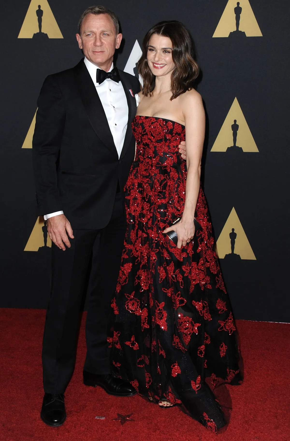 Rachel Weisz peeks out from beneath her black-and-red embroidered gown in delicate open-toe heels while posing beside husband Daniel Craig, whose classic black tuxedo emphasizes their noticeable 4¼-inch height difference on the red carpet