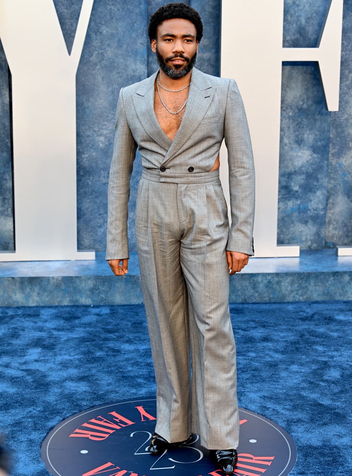 Donald Glover turned heads at the 2023 Vanity Fair Oscar Party in Beverly Hills, wearing a daring gray double-breasted jumpsuit with side cutouts and wide-leg tailoring, accessorized with layered Tiffany Co. necklaces and glossy black shoes