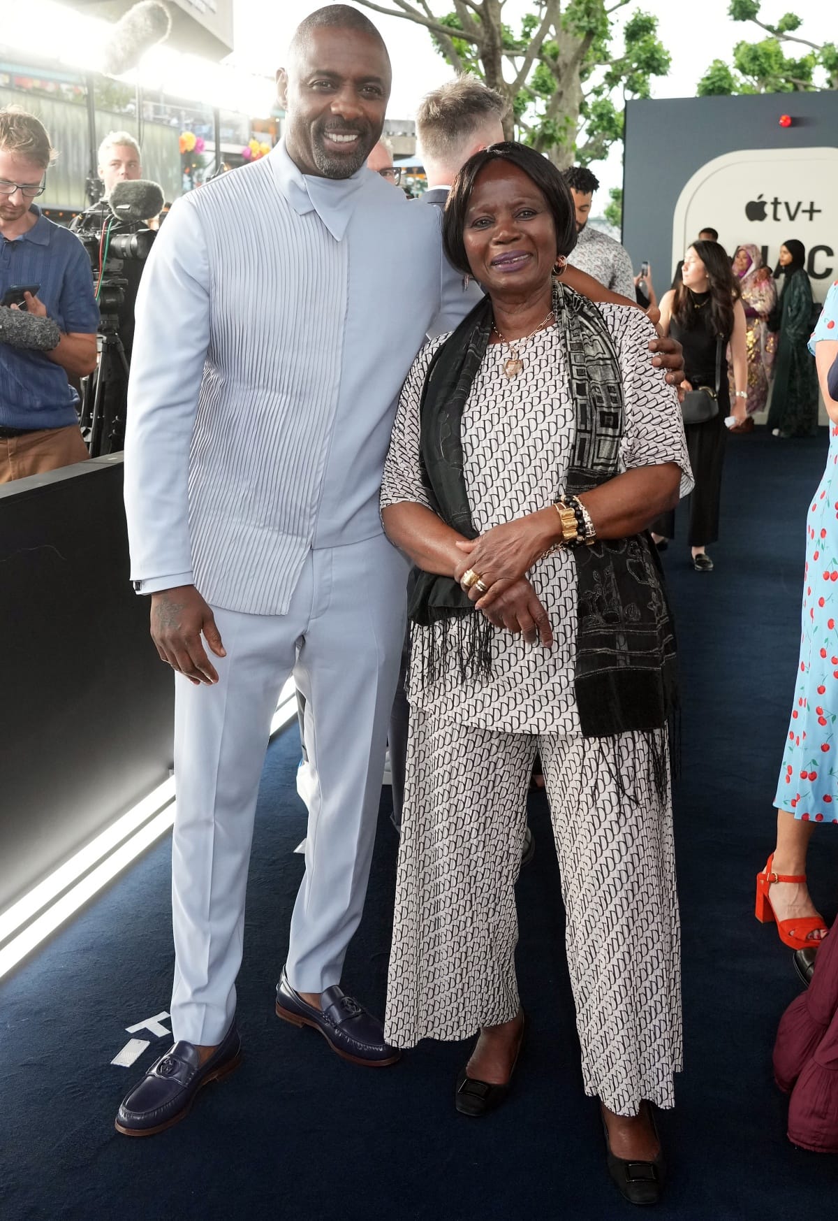 Idris Elba, dressed in a tailored pale blue suit with navy leather loafers, poses with his mother Eve Elba—who paired her patterned ensemble with classic black square-toe flats—at the world premiere of Hijack at BFI Southbank in London on June 27, 2023