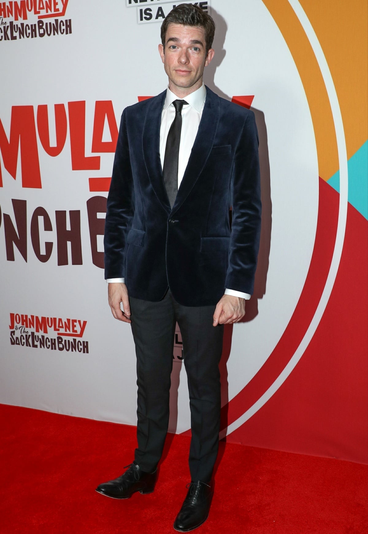 John Mulaney looked sharp at the New York City screening of John Mulaney The Sack Lunch Bunch, wearing a navy velvet blazer over a crisp white shirt, black tie, tailored trousers, and polished black dress shoes