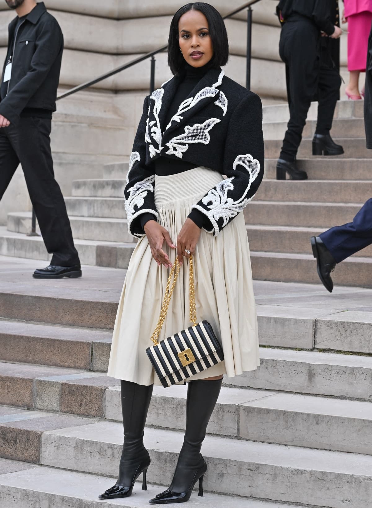 Sabrina Dhowre Elba attends the Schiaparelli Haute Couture Fall/Winter 2023/2024 show during Paris Fashion Week on July 3, 2023, in Paris, France, wearing a black-and-white embroidered cropped jacket, pleated cream skirt, and sleek pointed-toe knee-high boots, finished with a bold striped chain-handle handbag