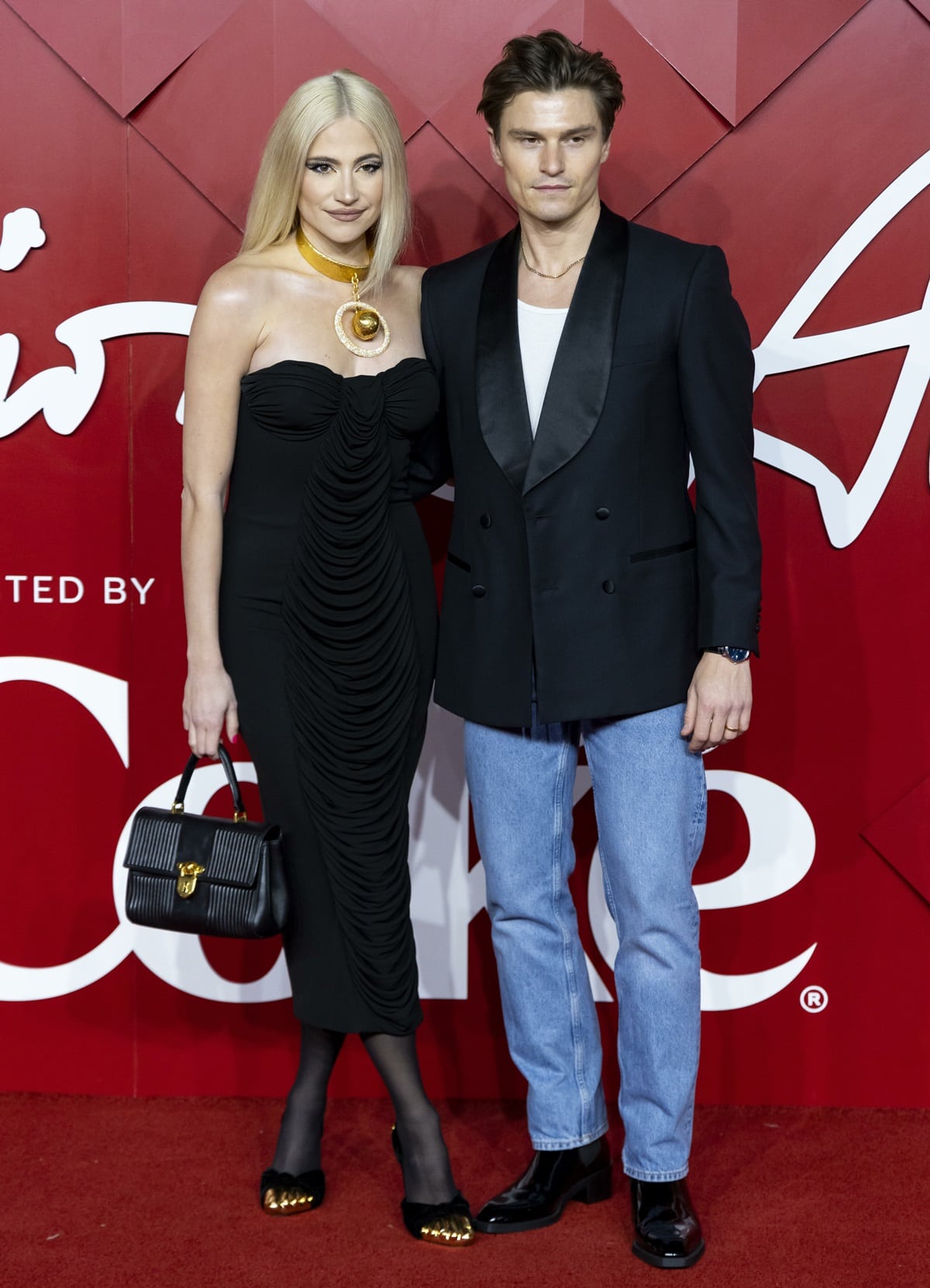 Pixie Lott stuns in an elegant black strapless dress, accessorized with a bold gold statement necklace and matching metallic-tipped shoes, while posing with husband Oliver Cheshire