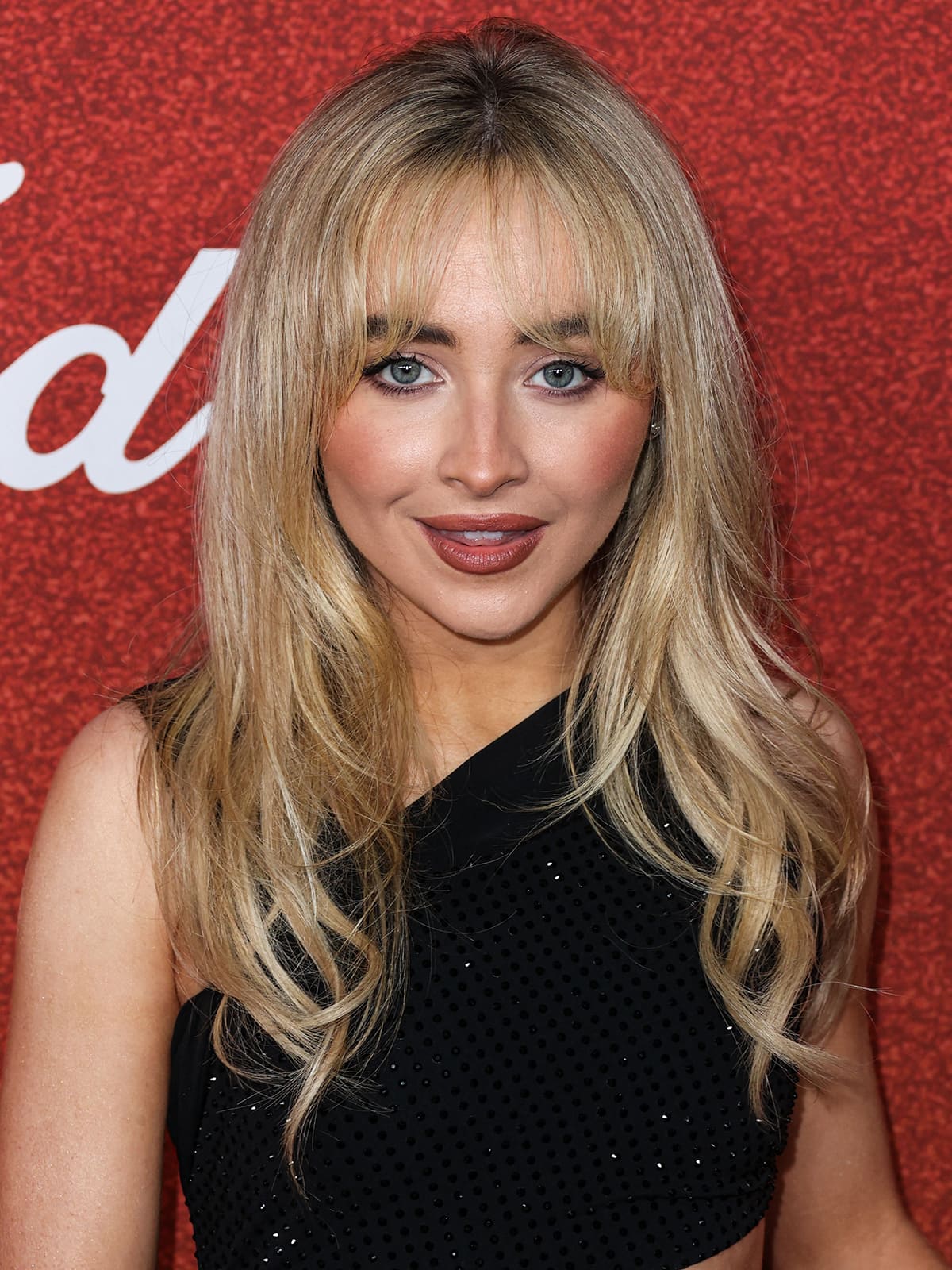 Sabrina Carpenter highlights her features with mascara, heavy blush, and berry lipstick and wears her signature blonde hair down in waves with her bangs covering her eyebrows