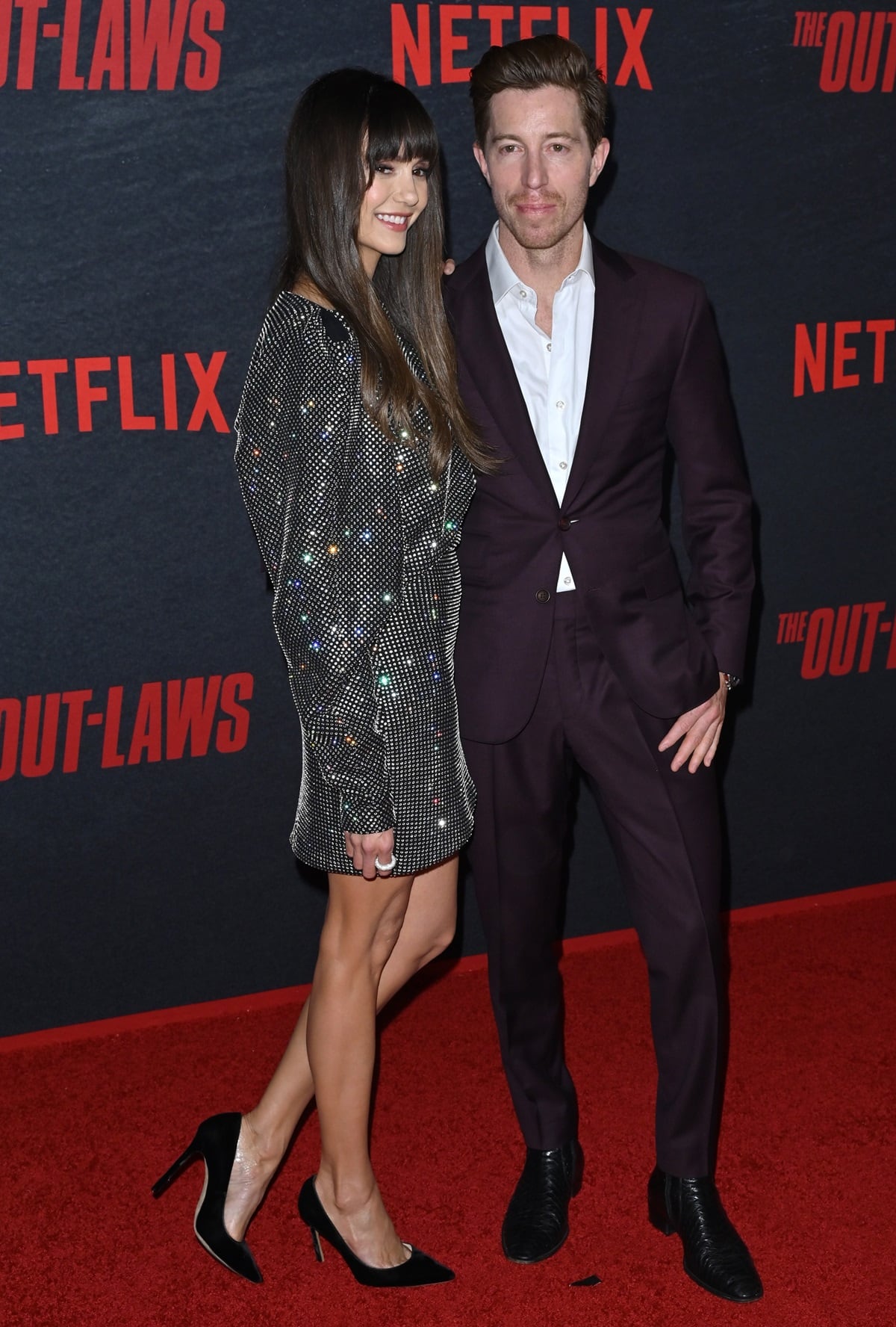 Nina Dobrev dazzles in a sparkling mini dress and black pumps, standing alongside Shaun White in a sharp burgundy suit at The Out-Laws premiere