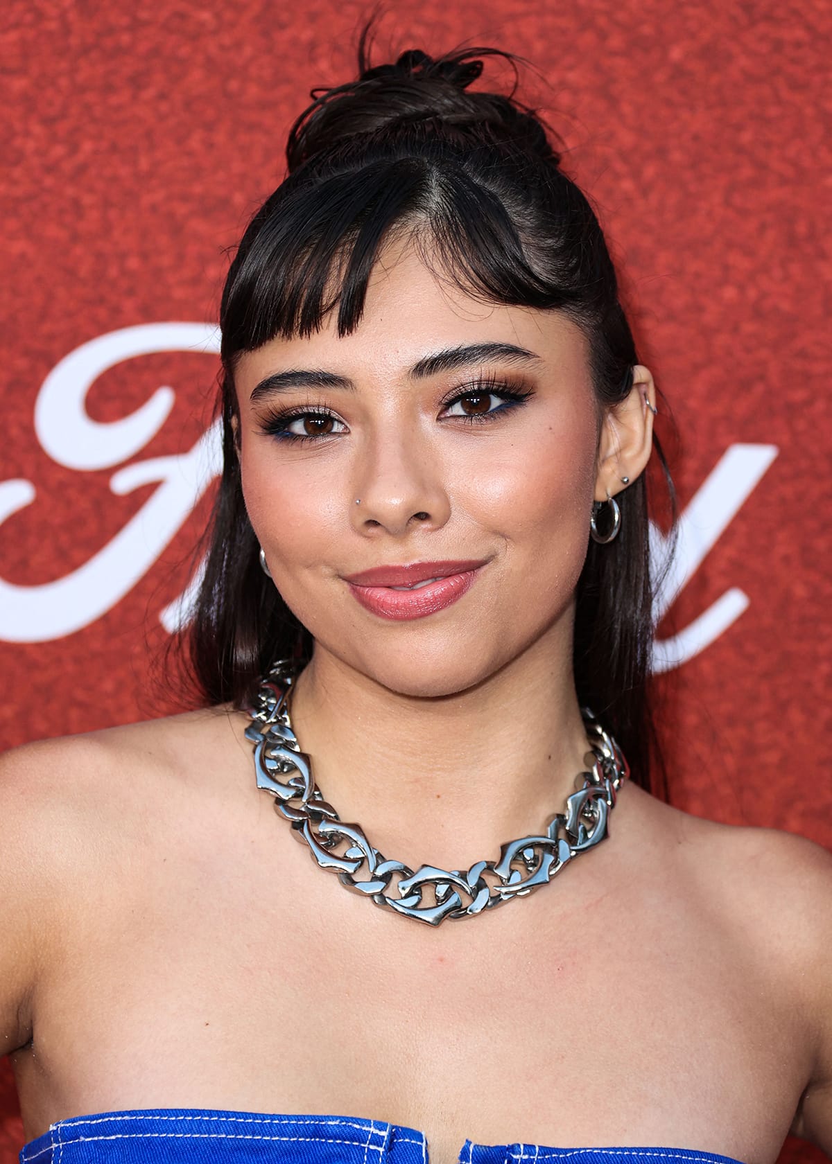 Xochitl Gomez wearing a chunky silver chain necklace, styling her hair in a chic half-up bun, and completing her glam with brown smoky eyeshadow, blush, and pink lipstick