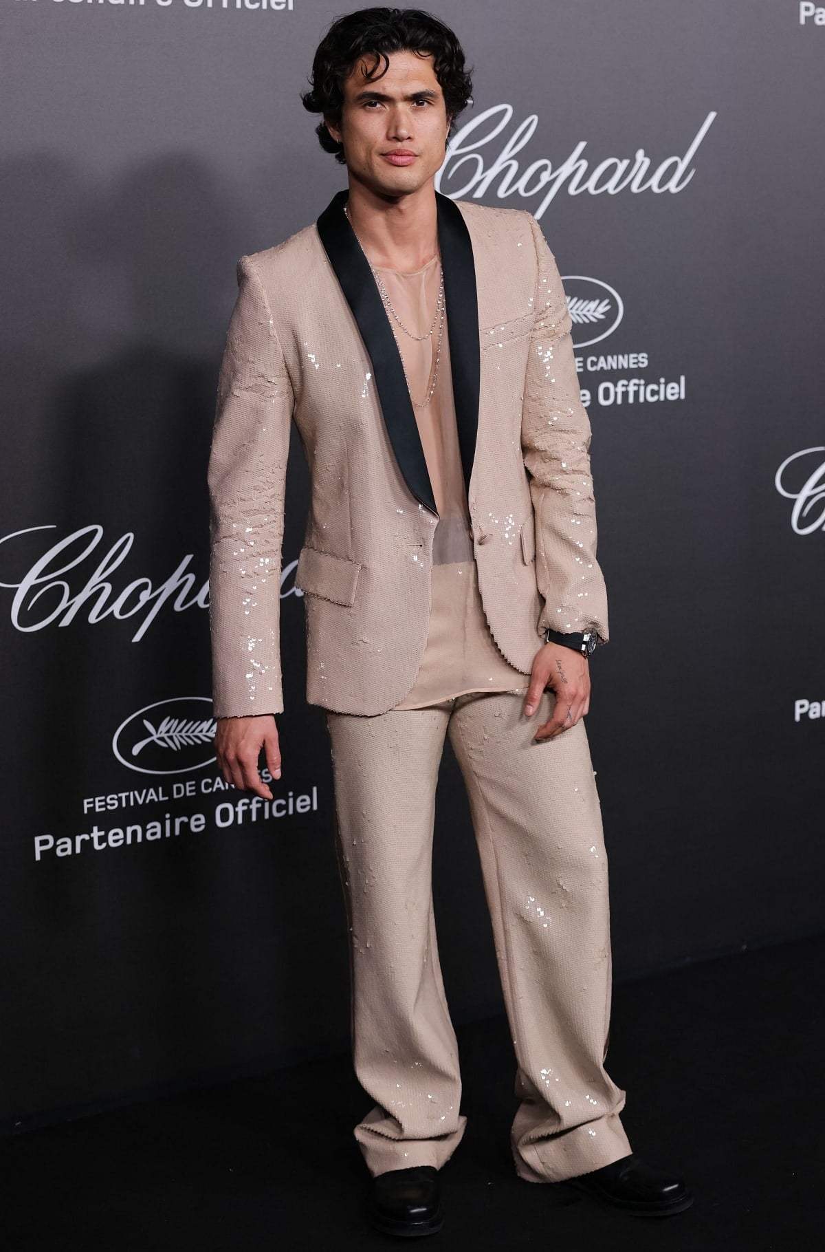 Styled by Jordan Dorso, Charles Melton wears a Valentino Spring 2023 sequined beige suit paired with a sheer top, layered chains, and sleek black leather boots at the Chopard event during Cannes