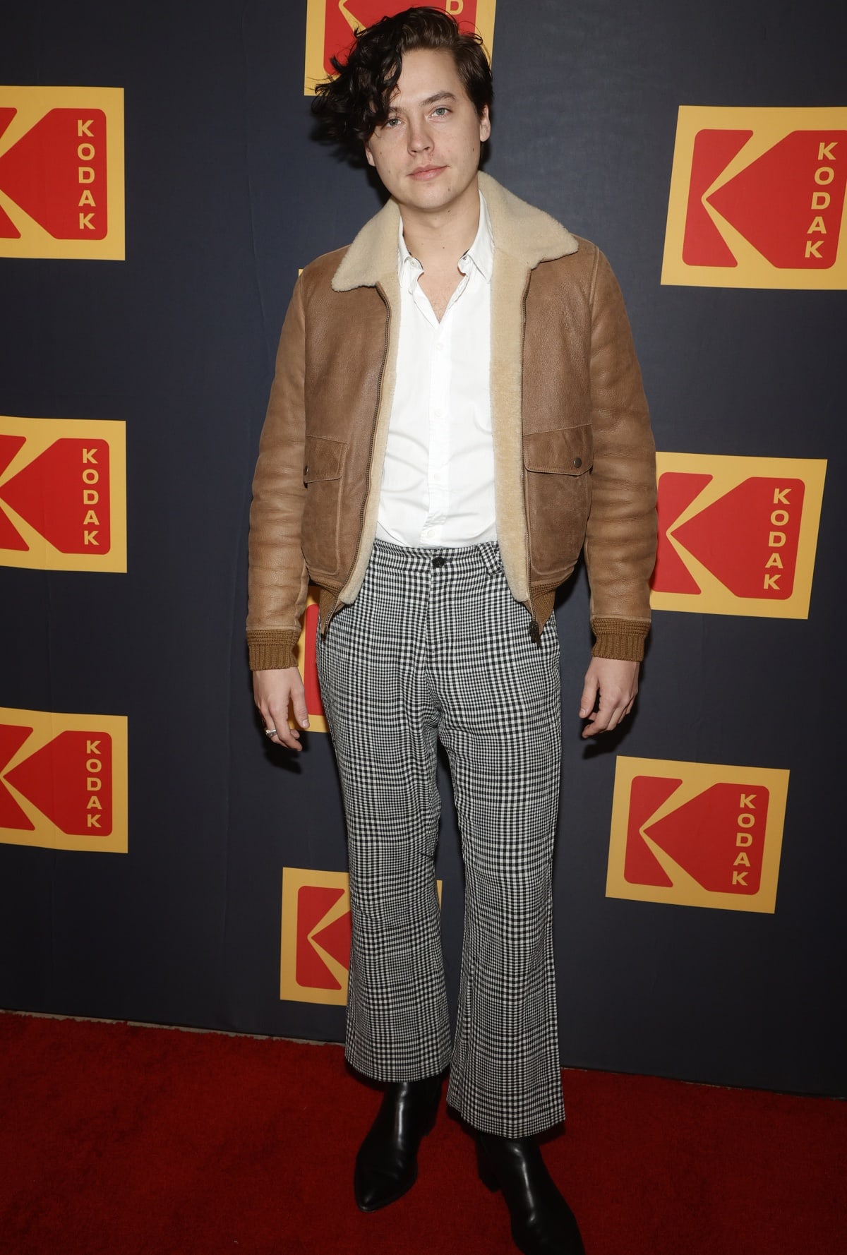 Cole Sprouse pairs his sharp black pointed-toe boots with retro-inspired plaid trousers, a crisp white shirt, and a cozy shearling bomber jacket at the Kodak event