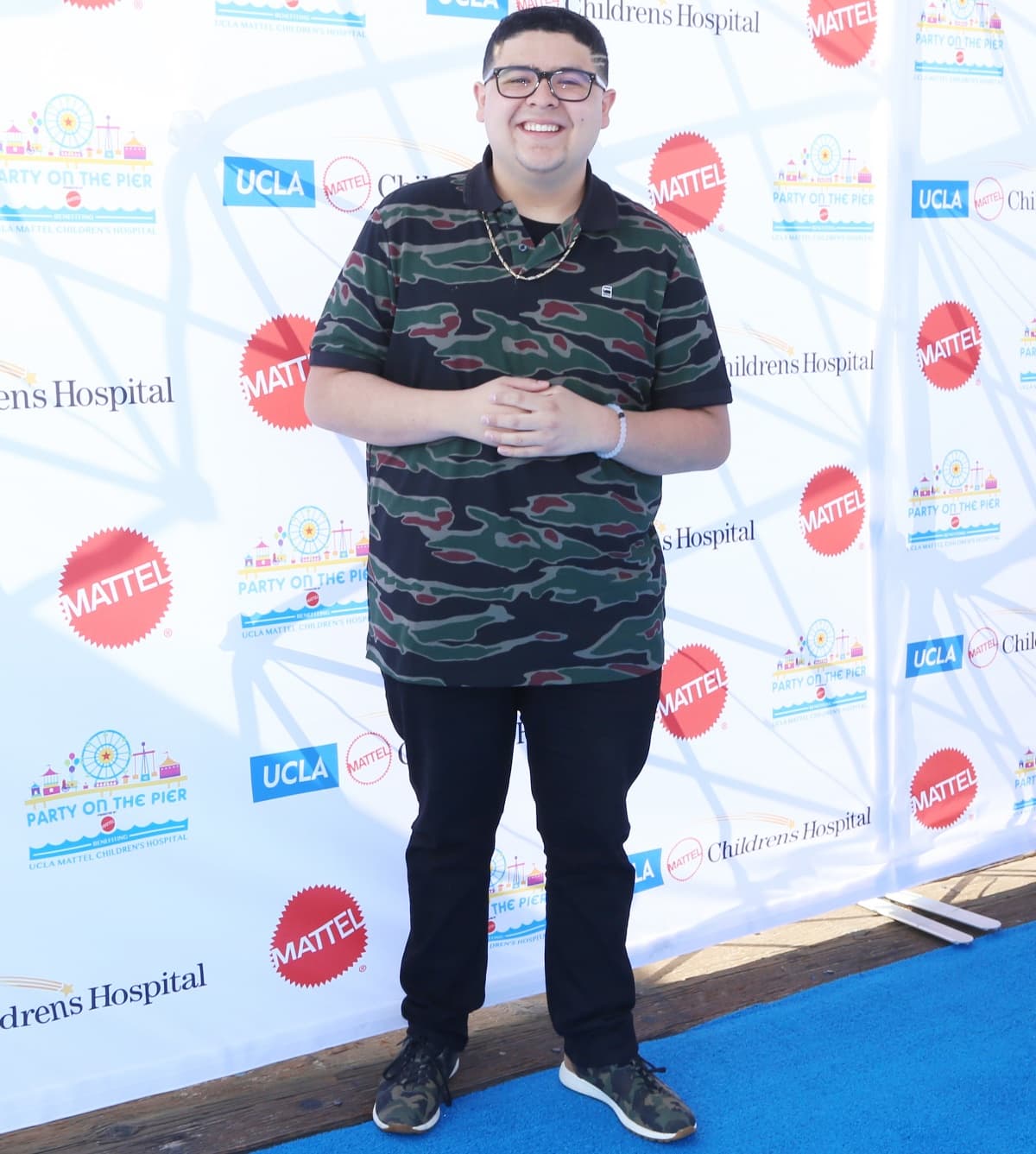Rico Rodriguez keeps it casual and coordinated in a camo-print polo shirt and matching sneakers, pairing the look with black pants and gold accessories for a playful, street-style vibe