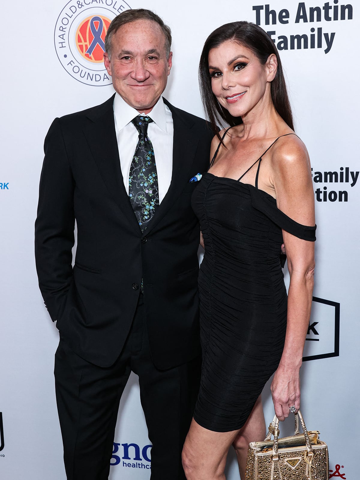 Heather Dubrow stuns in a ruched off-the-shoulder black mini dress paired with a gold-studded Prada handbag as she poses with her husband, Dr. Terry Dubrow
