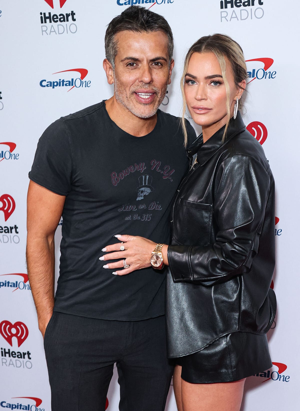 Teddi Mellencamp Arroyave steps out in a sleek black faux leather shirt-and-shorts set, accessorized with a bold gold watch and hoop earrings, as she poses with husband Edwin Arroyave at the iHeartRadio event