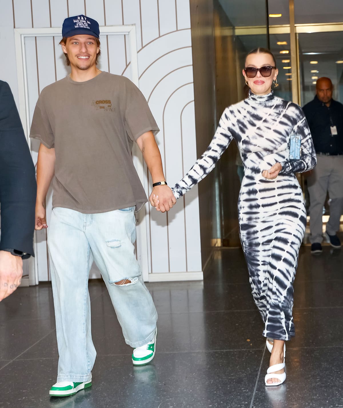 Millie Bobby Brown steps out in a figure-skimming tie-dye Joseph dress paired with white Steve Madden puff-strap heels, while Jake Bongiovi keeps things casual in oversized denim, a taupe tee, and green-and-white sneakers as the couple leave The Today Show in New York City following press for Nineteen Steps on September 14, 2023