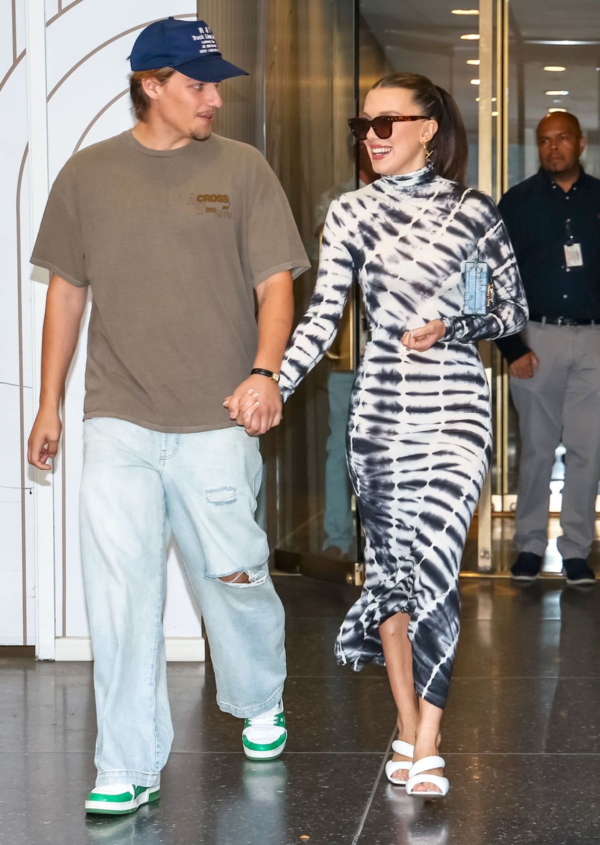 Millie Bobby Brown stuns in a chic black-and-white tie-dye maxi dress paired with sleek white sandals, effortlessly complementing Jake Bongiovi's casual look
