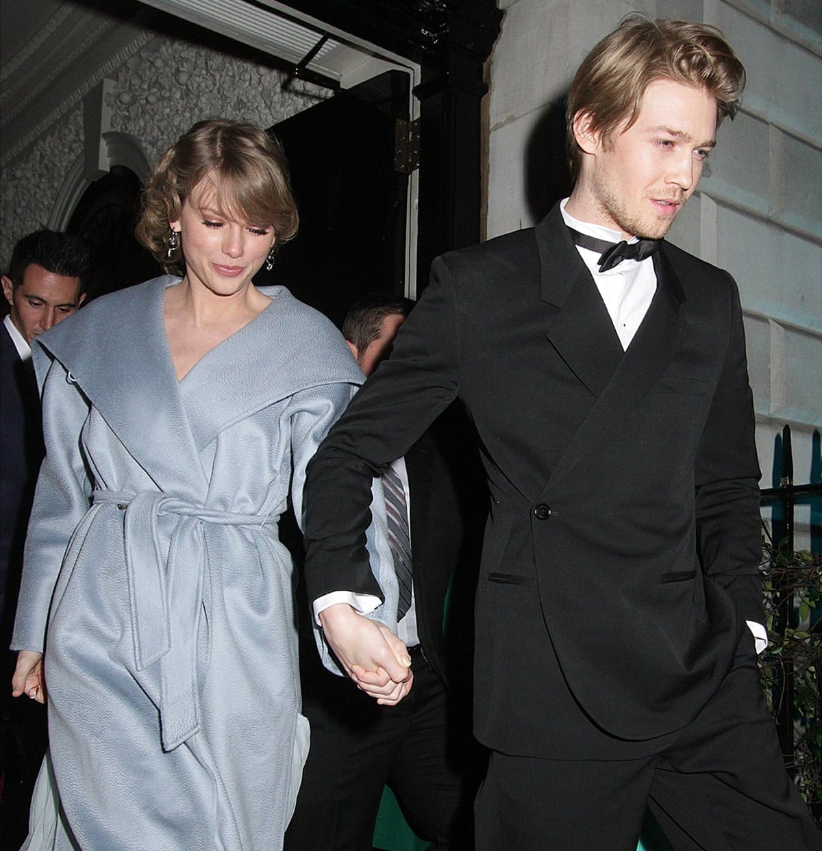 Taylor Swift and Joe Alwyn hold hands as they leave an event together, looking elegant in a light blue coat and black tuxedo
