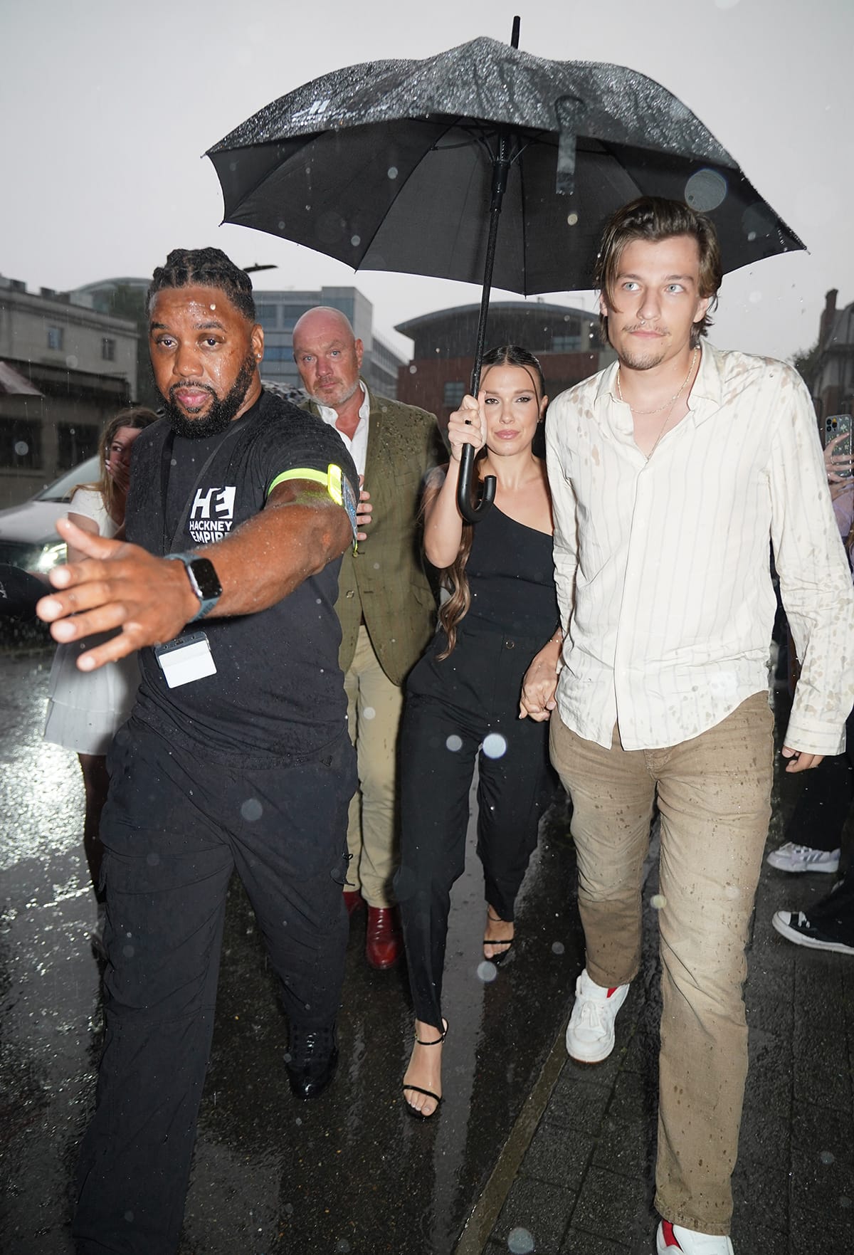 Millie Bobby Brown braves the London rain in a sleek black one-shoulder ensemble styled with delicate strappy heels, while Jake Bongiovi complements her understated evening look in a striped cream shirt, khaki trousers, and white sneakers at the Nineteen Steps book launch at Hackney Empire on September 12, 2023
