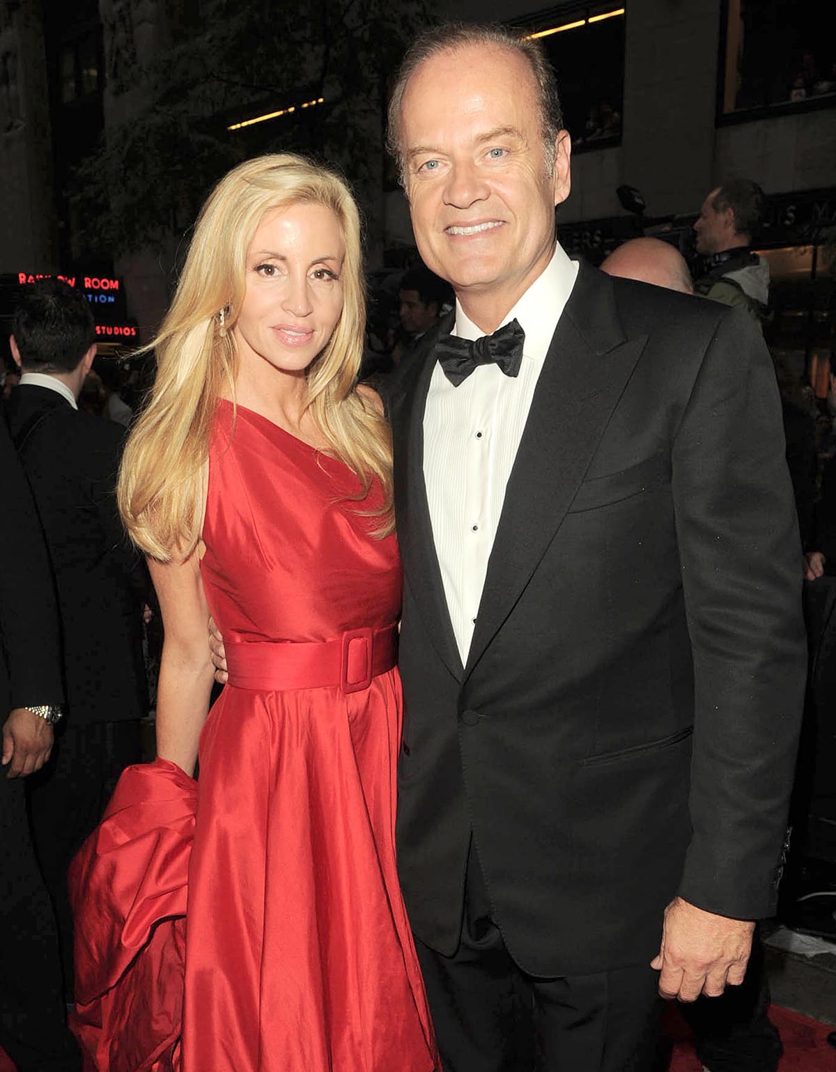 Camille Grammer and then-husband Kelsey Grammer arrive in elegant style—Camille glowing in a classic belted red satin gown with a voluminous train, and Kelsey in a sharp black tux—at a formal red carpet event