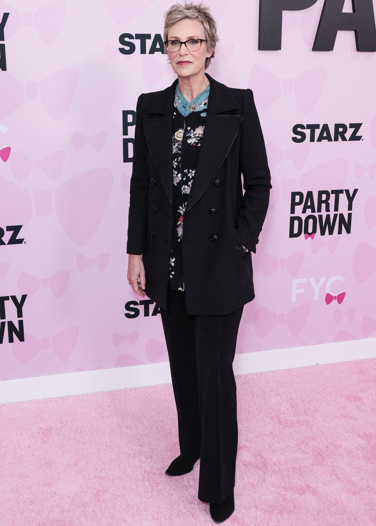 Jane Lynch embraced understated elegance in a floral blouse layered under a tailored black coat and wide-leg trousers, finishing the look with sleek black pointed-toe shoes at the Party Down FYC event