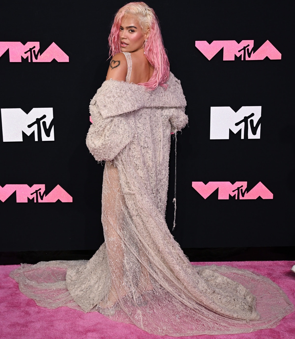 Karol G wearing a floor-length distressed coat to add a dramatic twist to her sleeveless mermaid dress
