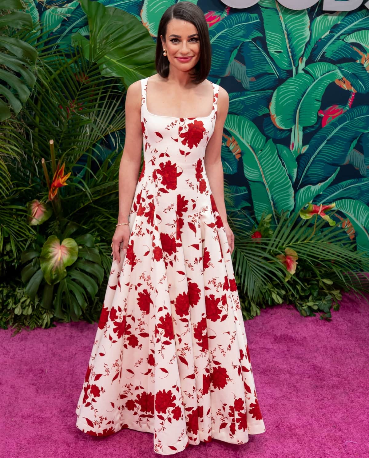 Lea Michele exuded timeless elegance in a white floor-length gown adorned with bold red floral prints, gracefully concealing her shoes beneath the flowing hemline