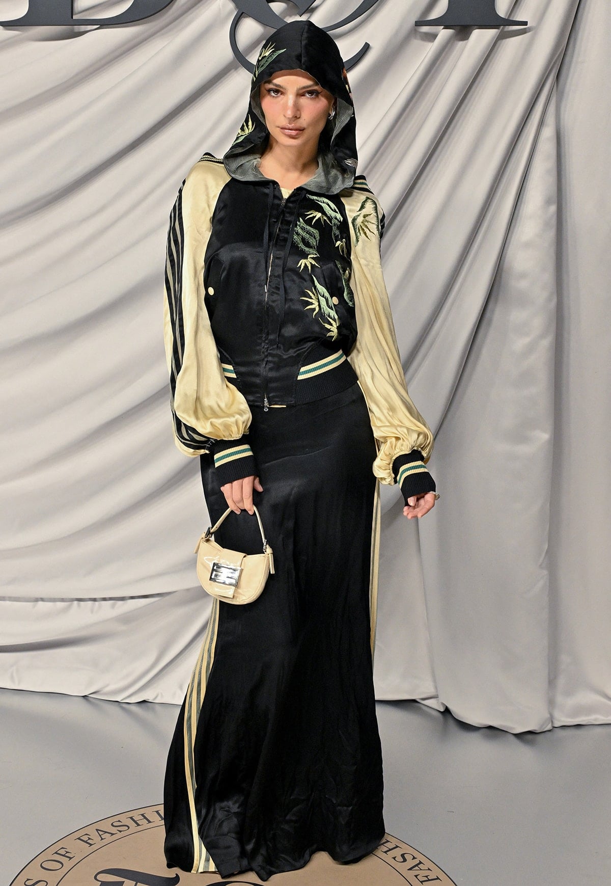 Emily Ratajkowski wears a black dress underneath a black satin bomber jacket with an embroidered hood, puffy sleeves, and striped detailing