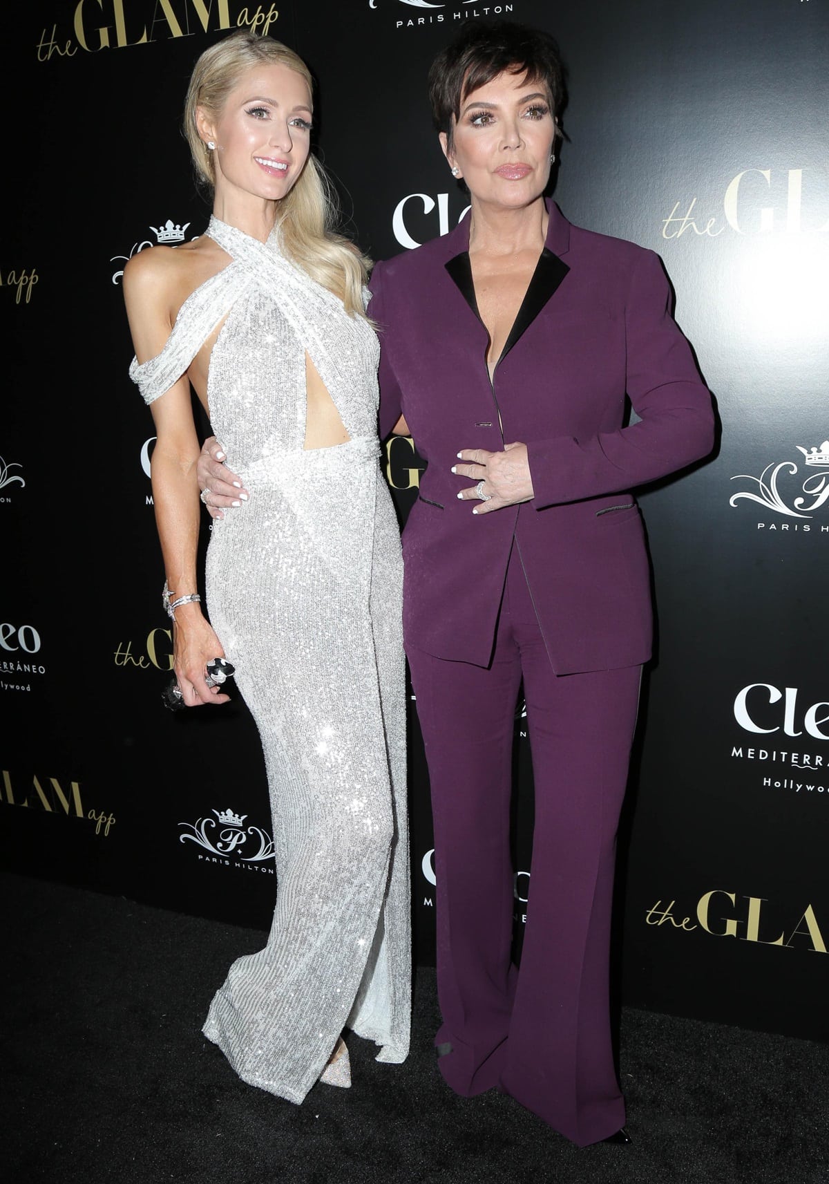 Kris Jenner looks polished in a tailored plum pantsuit while posing with Paris Hilton, who sparkles in a silver cut-out gown, at the launch of The Glam App in Hollywood