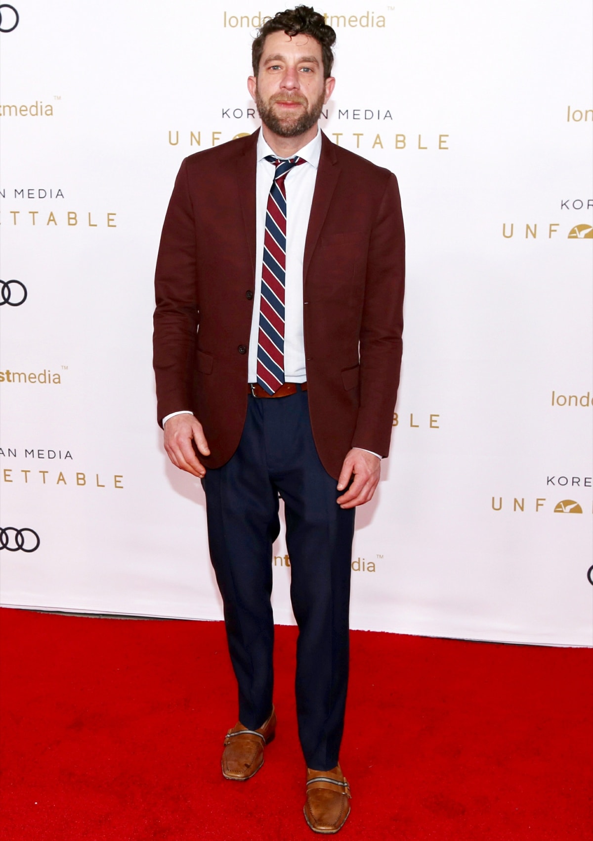 Elliott Yamin keeps it classic on the red carpet in a burgundy blazer, striped tie, and navy slacks, finishing the look with tan suede loafers for a touch of relaxed polish