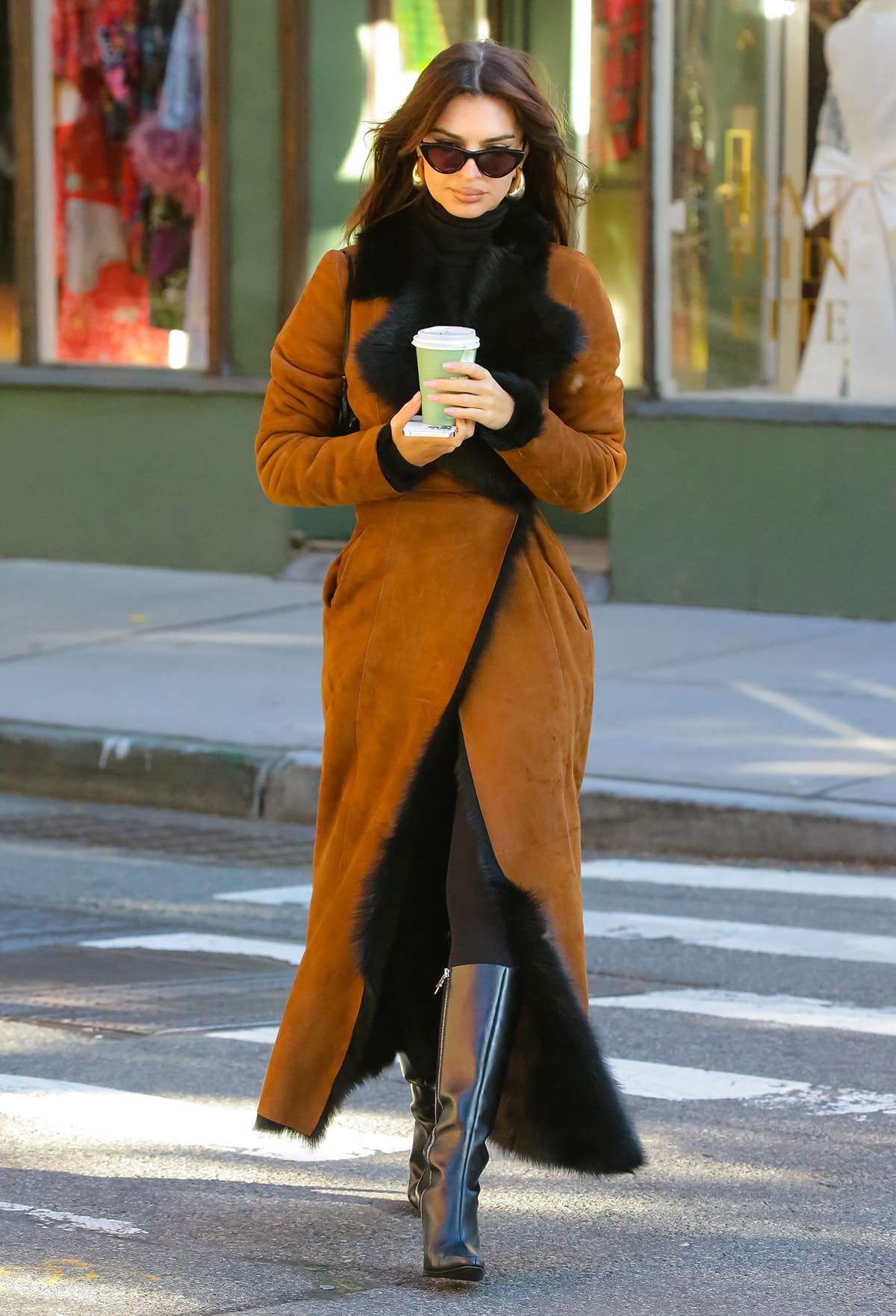 Emily Ratajkowski cuts an elegant figure in a cognac suede shearling coat layered over sleek black knee-high boots while crossing a Manhattan street on November 15, 2023