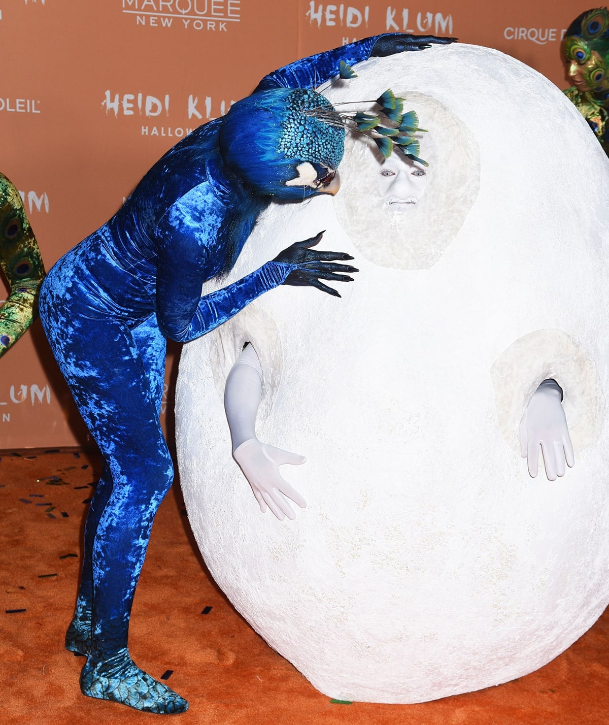 Heidi Klum playfully interacts with husband Tom Kaulitz, who appeared as a giant peacock egg, while wearing textured blue bird-feet boot covers over heels