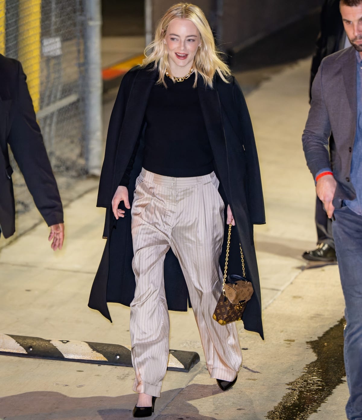 Emma Stone steps out in champagne pinstripe trousers and a draped navy coat, finishing the polished look with black pointed-toe heels and a monogram Louis Vuitton bag