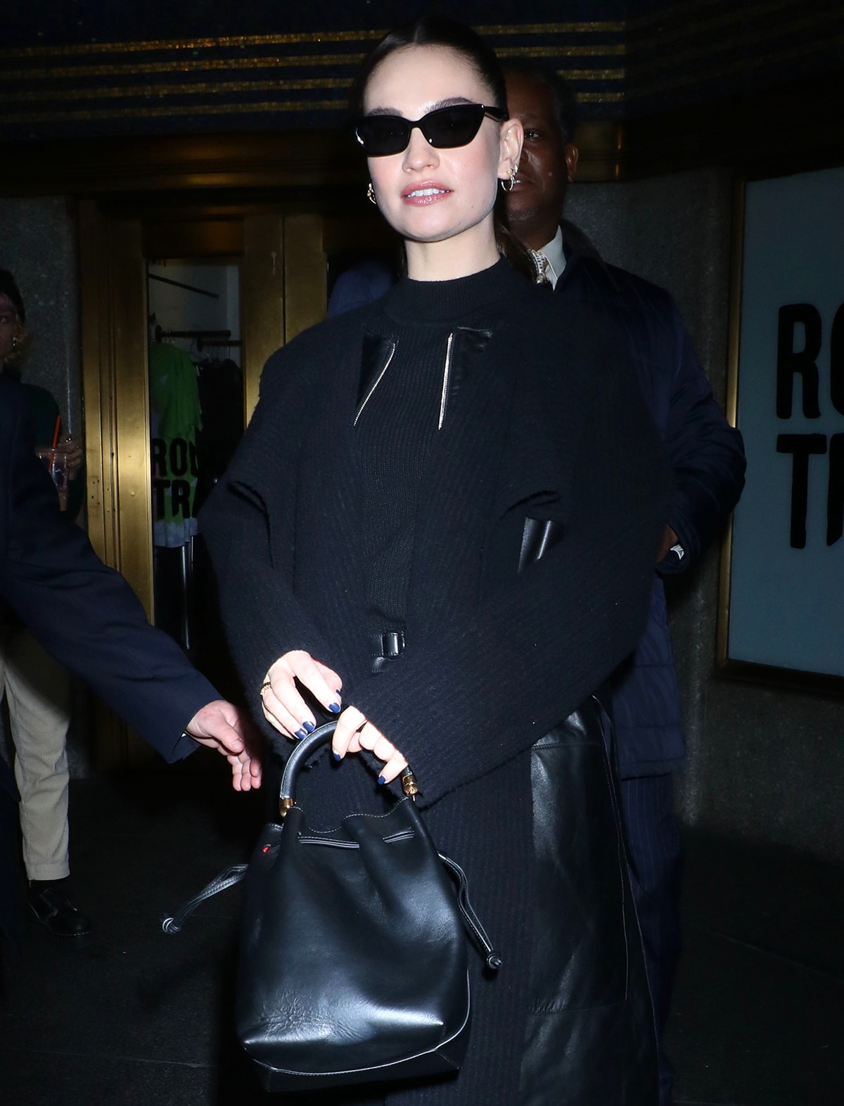 Lily James accessorizes her monochromatic black outfit with Oscar Frank sunnies and a black Mansur Gavriel bucket bag