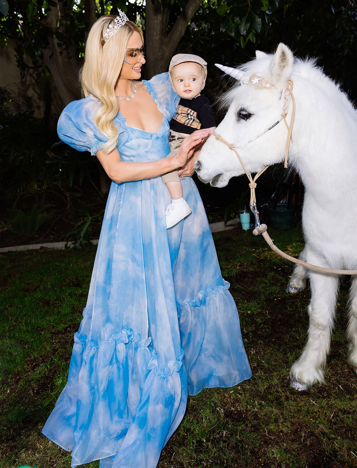 Paris Hilton, wearing her cloud-printed Selkie princess gown, smiles as she and son Phoenix gently pet a white pony dressed as a unicorn from Itty Bitty Petting Zoo