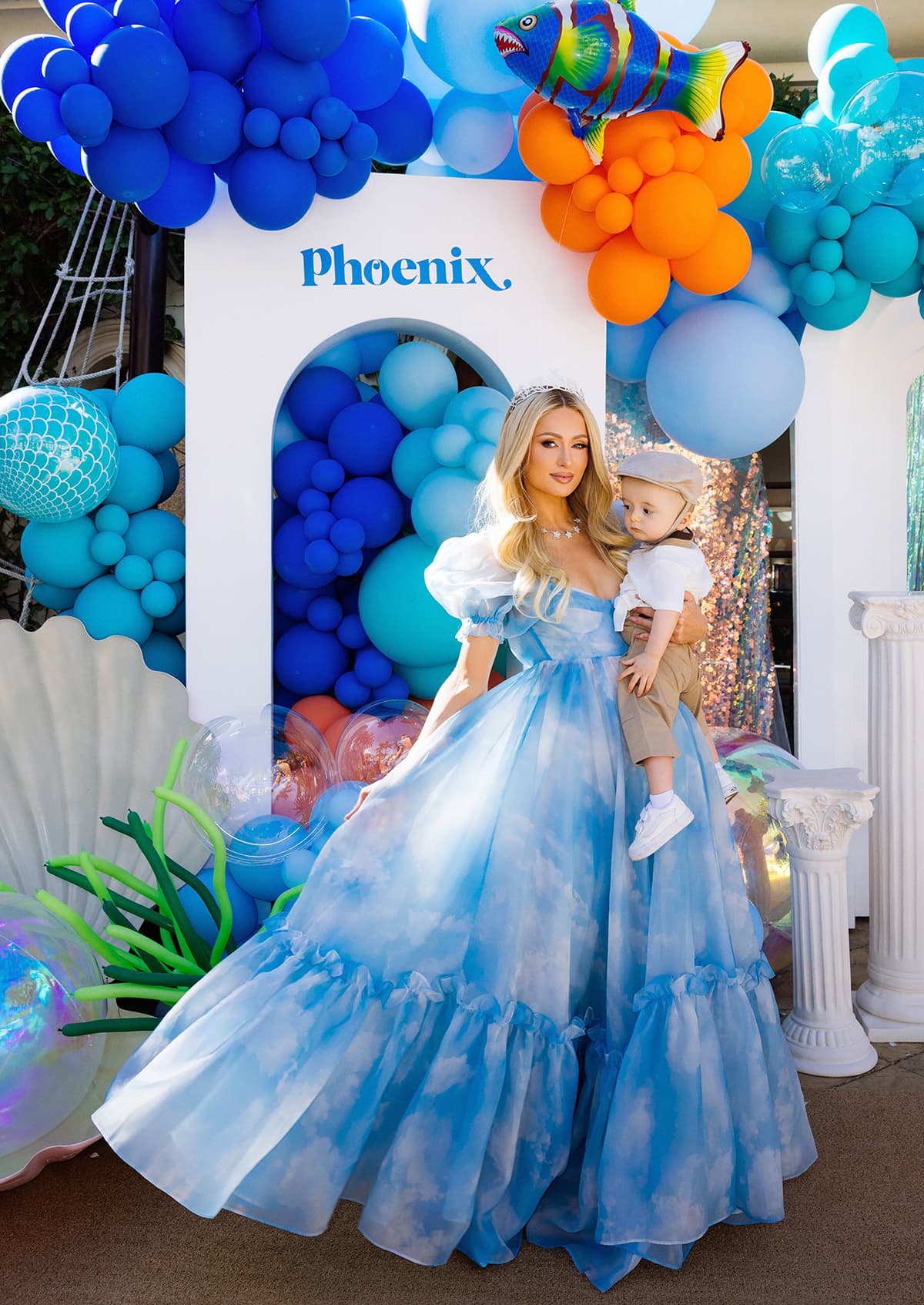 Paris Hilton carries her son Phoenix—dressed in a white Fendi tee, tan pants, and tiny white sneakers—while posing in front of a vibrant, ocean-themed balloon backdrop created by Balloon and Paper