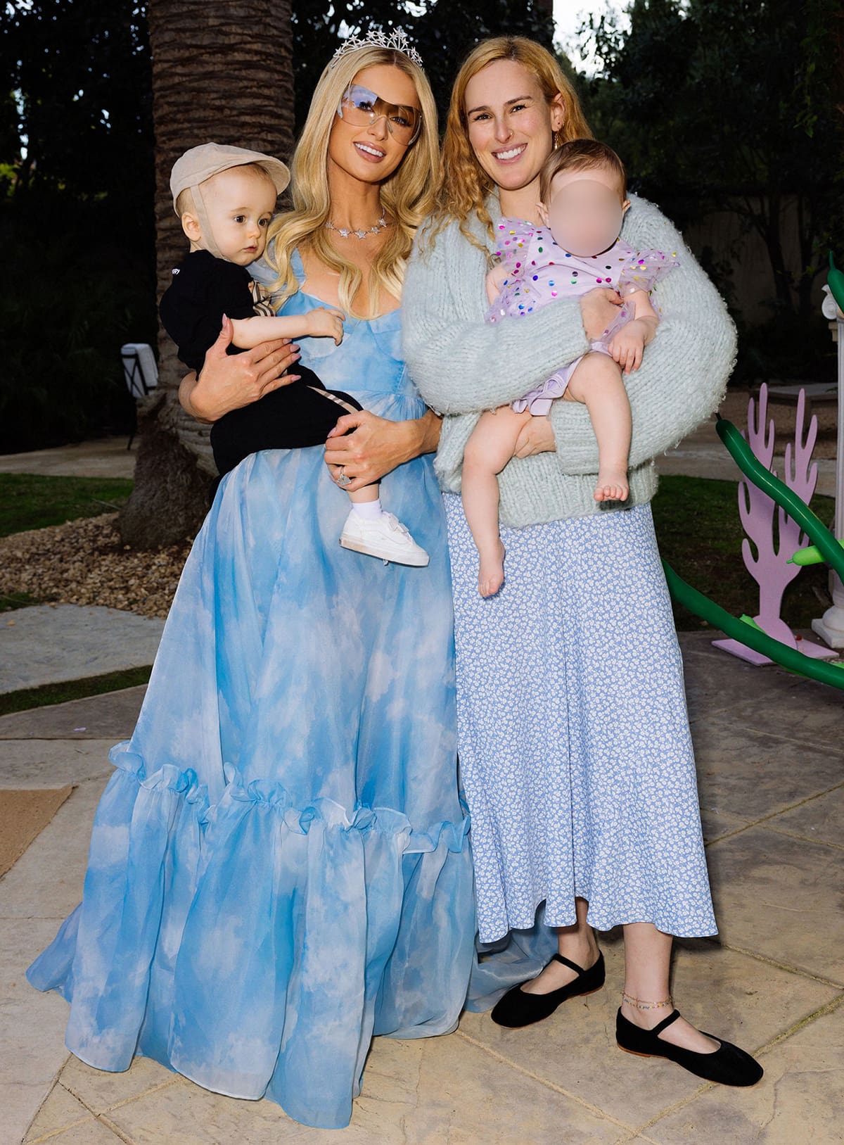 Paris Hilton poses with Rumer Willis, who wears a cozy mint cardigan, a blue floral midi skirt, and classic black ballet flats while holding her daughter, at Phoenix’s “Sliving Under the Sea” birthday celebration