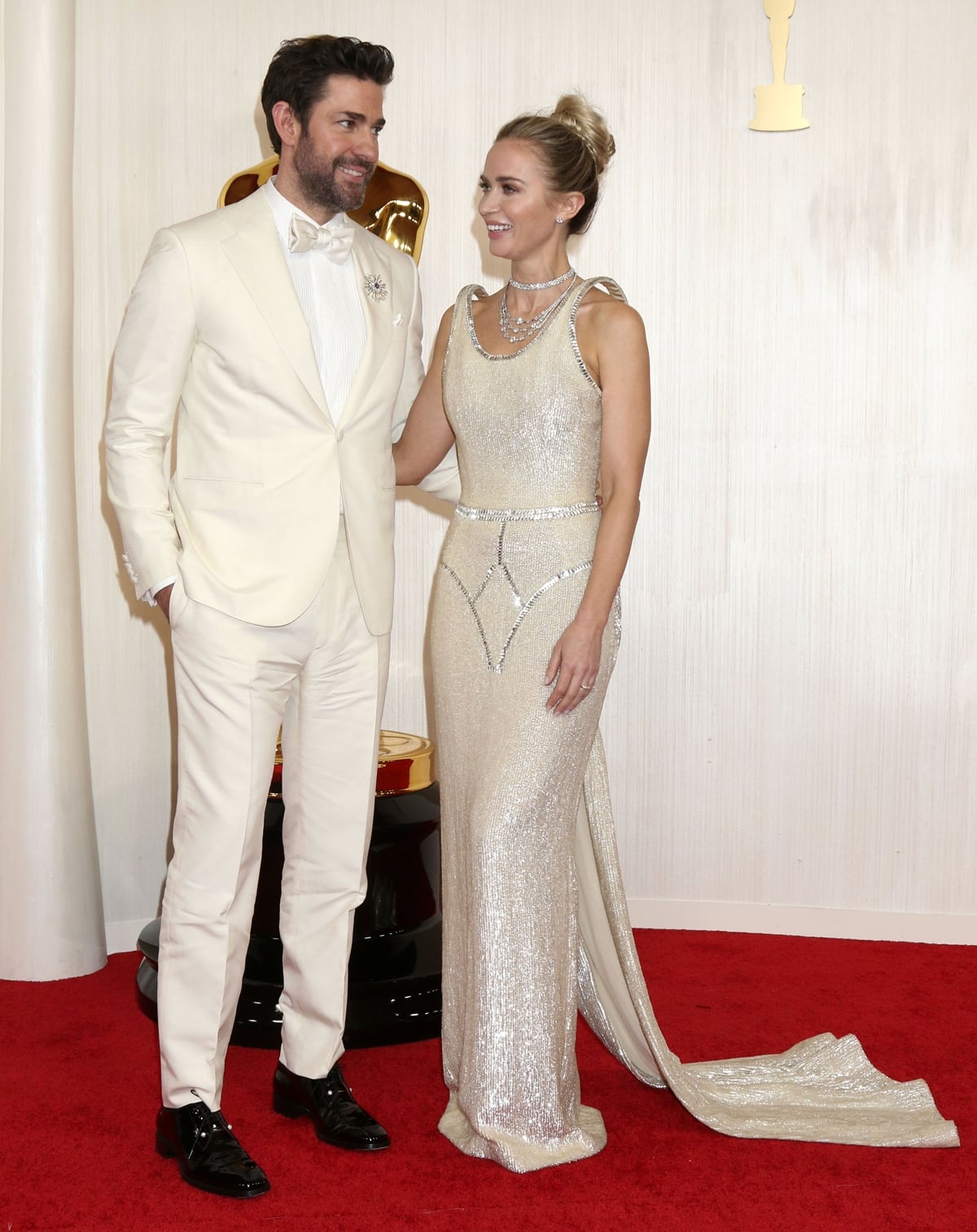Emily Blunt, accompanied by her husband John Krasinski, makes a stunning entrance at the 2024 Academy Awards, dressed in a breathtaking gold Schiaparelli gown from the Spring/Summer 2024-2025 couture collection, featuring ethereal floating sleeves and clear crystal adornments, showcasing her statuesque elegance