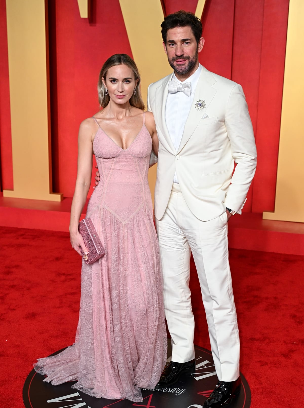 At the Vanity Fair Oscar Party, Emily Blunt switches to a custom-made pale pink lace gown by Dolce Gabbana, playfully nodding to the Barbie movie, with John Krasinski complementing her look in a classic cream tuxedo, exemplifying Hollywood glamour and sophistication