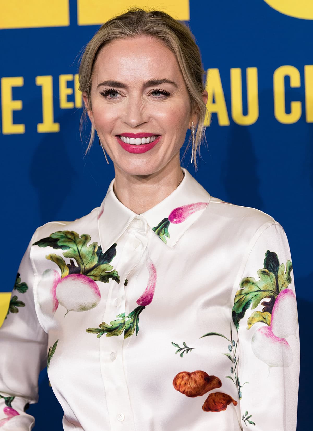 Emily Blunt beams on the red carpet in a satin Loewe blouse adorned with playful vegetable prints, paired with soft glam makeup and fuchsia lipstick