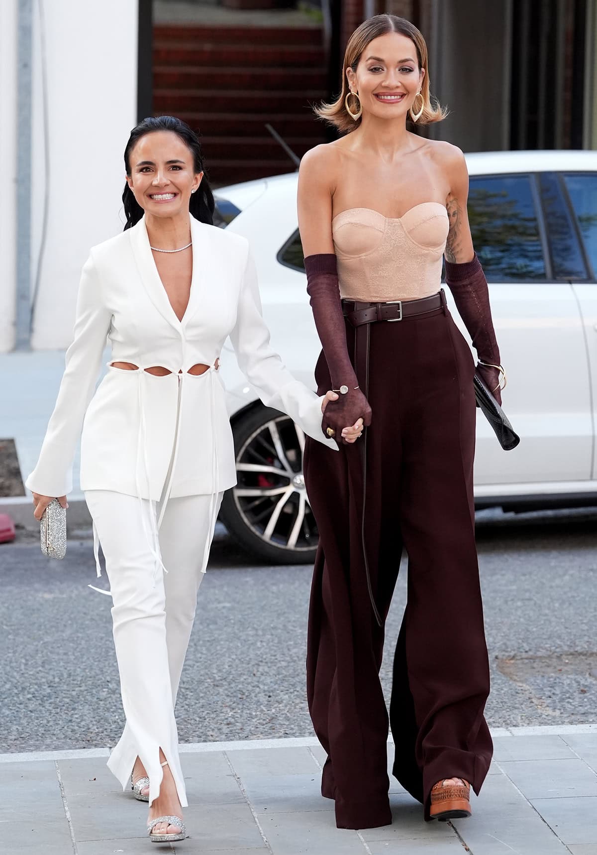 Anna Lahey, Vida Glow founder, arrives hand-in-hand with Rita Ora, pairing her white cutout suit with silver crystal-embellished sandals, while Ora grounds her corset-and-wide-leg look with chunky brown platform heels that add height and retro edge