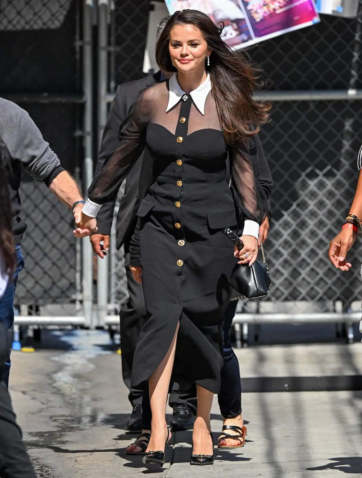 Selena Gomez exudes elegance in a black Versace dress featuring sheer sleeves, a sweetheart-style bodice, gold Medusa buttons, and crisp white collar and cuffs, perfectly paired with patent leather pumps