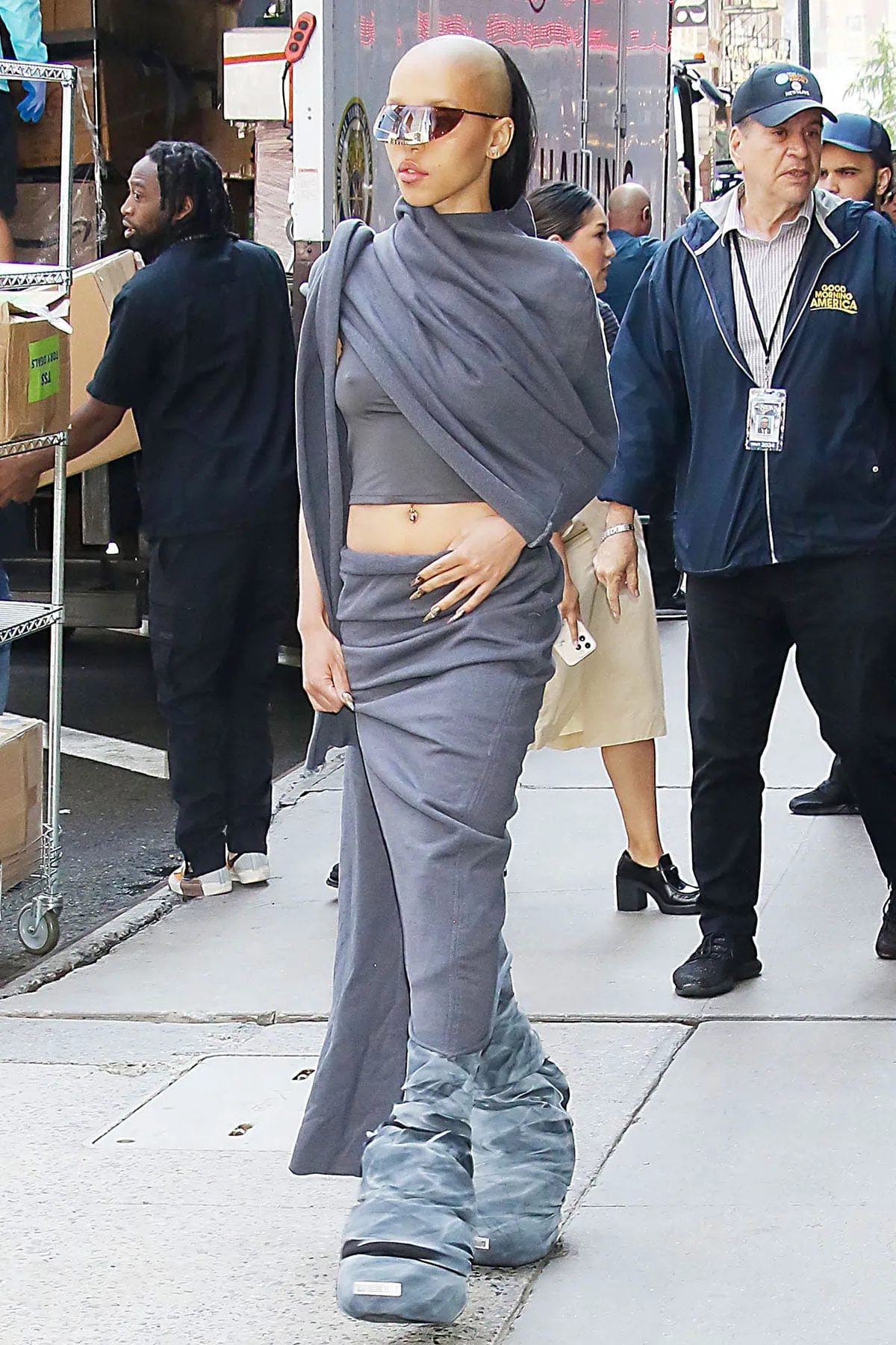 FKA Twigs leans into dystopian minimalism in an asymmetric gray Rombaut crop top and draped midi skirt, finishing the look with oversized, cloud-like boots that add a sculptural, otherworldly edge as she exits the Good Morning America studios
