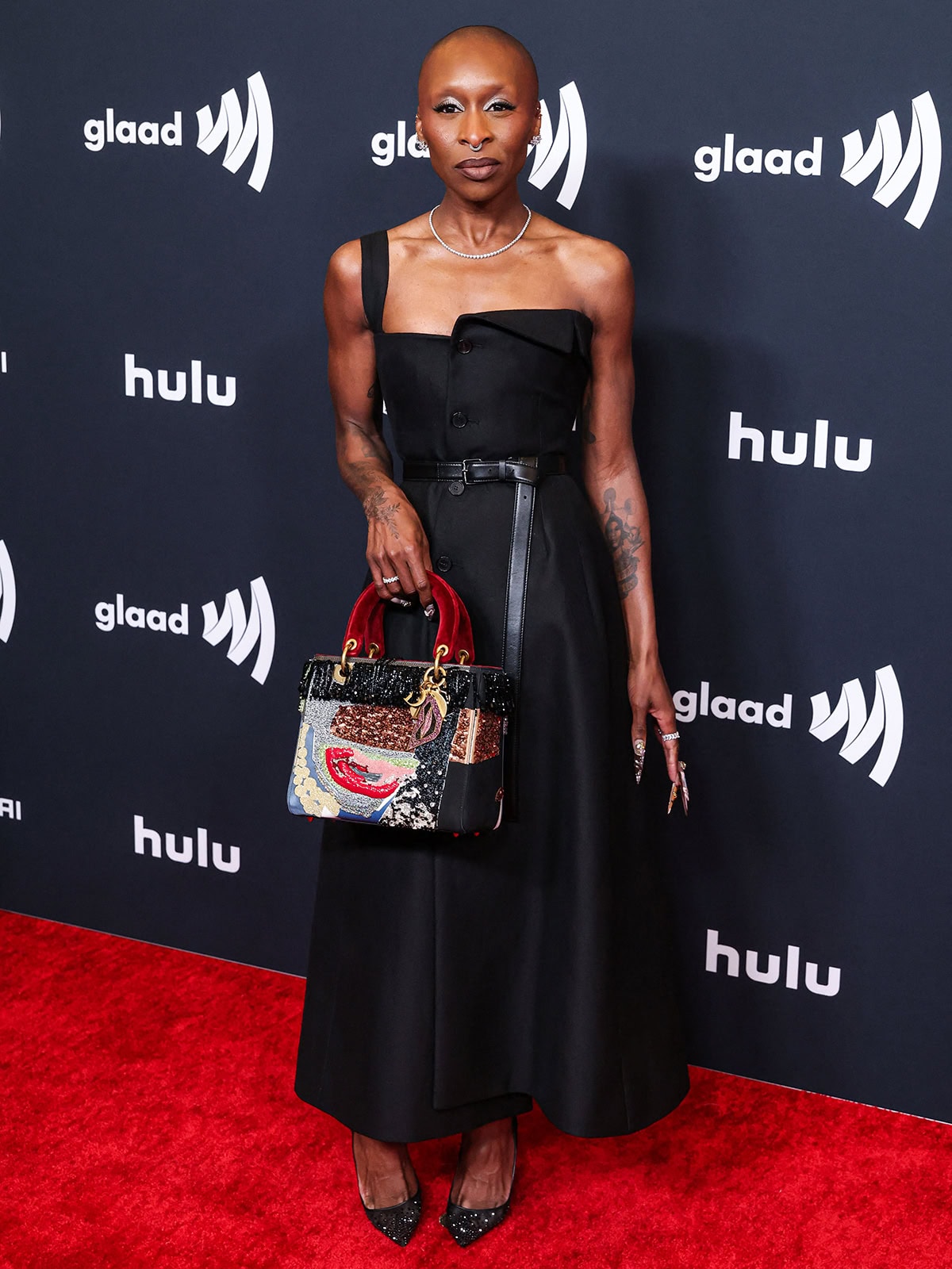 Cynthia Erivo embraces simplicity in a chic black dress by Dior featuring a one-shoulder strap, a folded collar at the neckline, front buttons, and a dangling belt