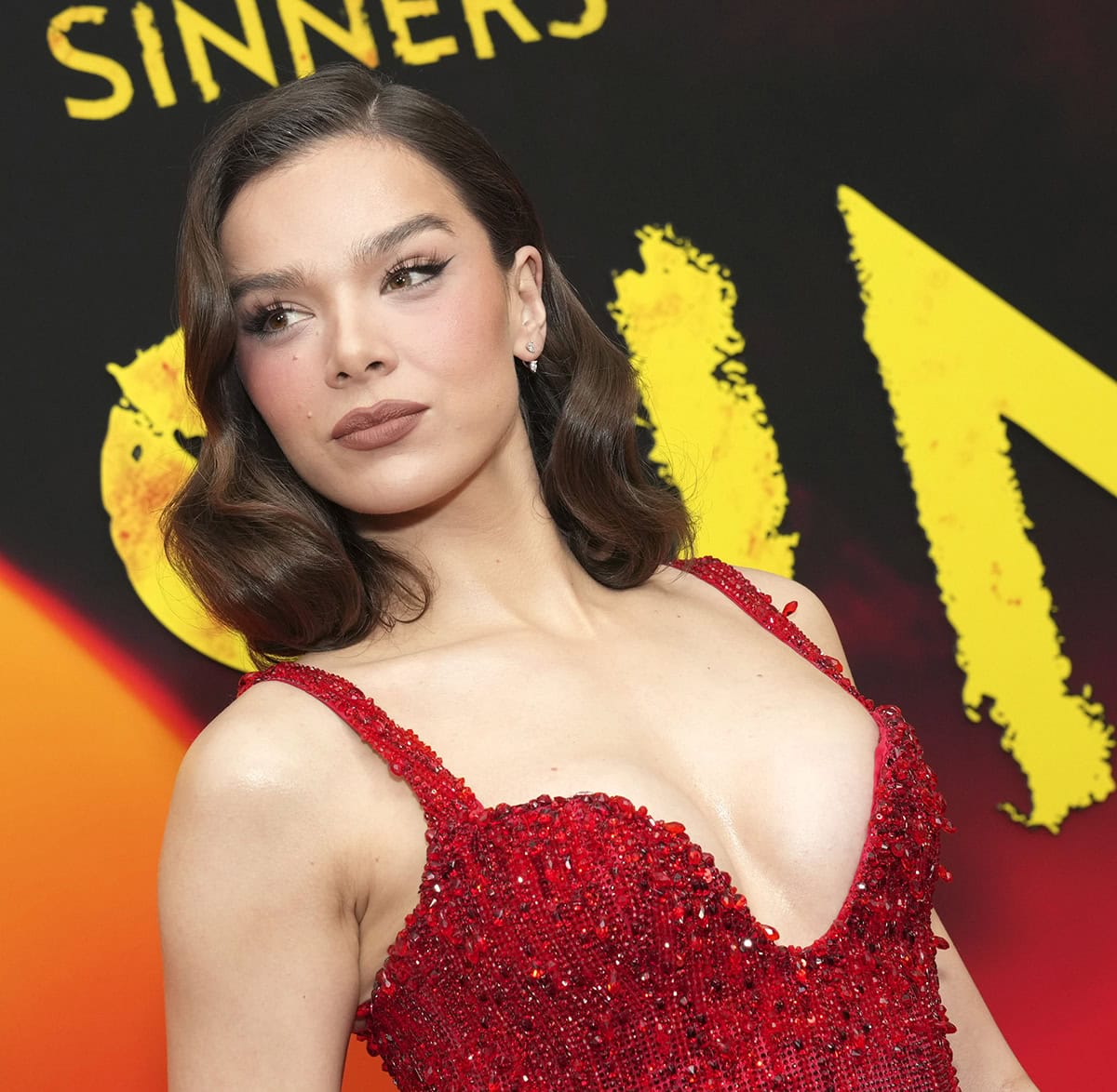Hailee Steinfeld styles her hair in Old Hollywood side-parted waves and wears neutral makeup with fluttery eyelashes, winged eyeliner, and nude lipstick