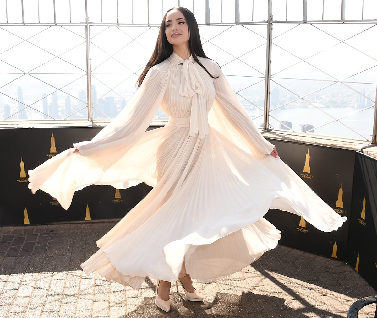 Sofia Carson twirls in an airy ivory chiffon Dolce Gabbana gown, featuring a pussy bow, a tonal belt, and delicate pleats, at the Empire State Building
