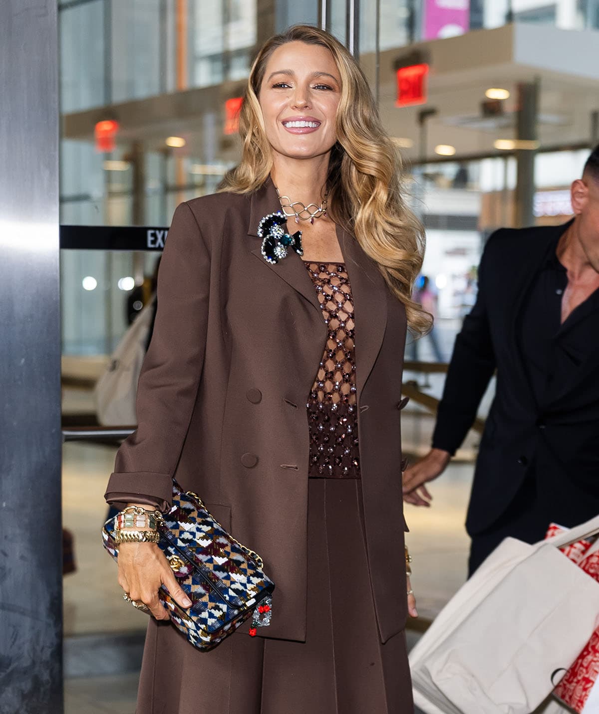 Blake Lively adds a playful twist to her refined skirt suit with a geometric gold necklace, stacked chunky bangles, statement rings, a bejeweled brooch, and a multicolored Chanel flap bag