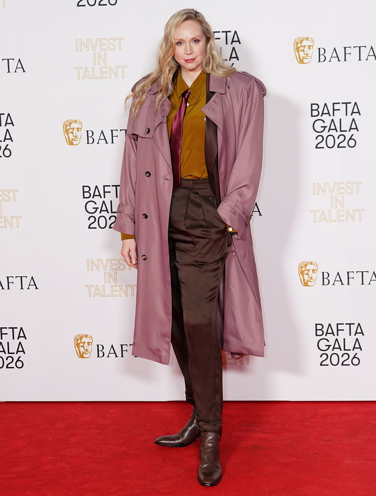Gwendoline Christie embraces a colorful, androgynous look, mixing a mustard button-down shirt with a burgundy silk tie, brown pants, and a rose-colored trench coat